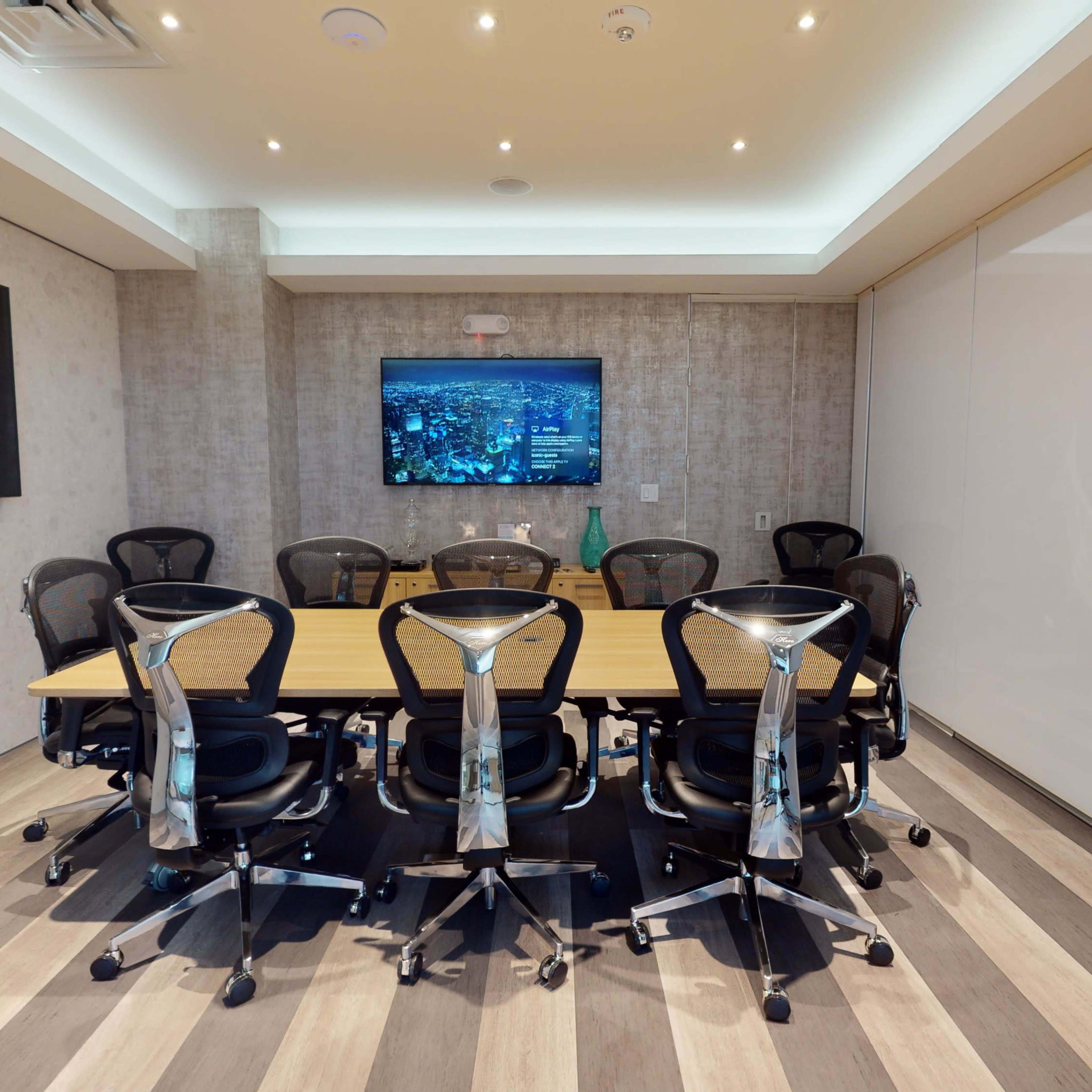 A rectangular conference room features a large table surrounded by ergonomic chairs, with a wall-mounted screen displaying visuals.