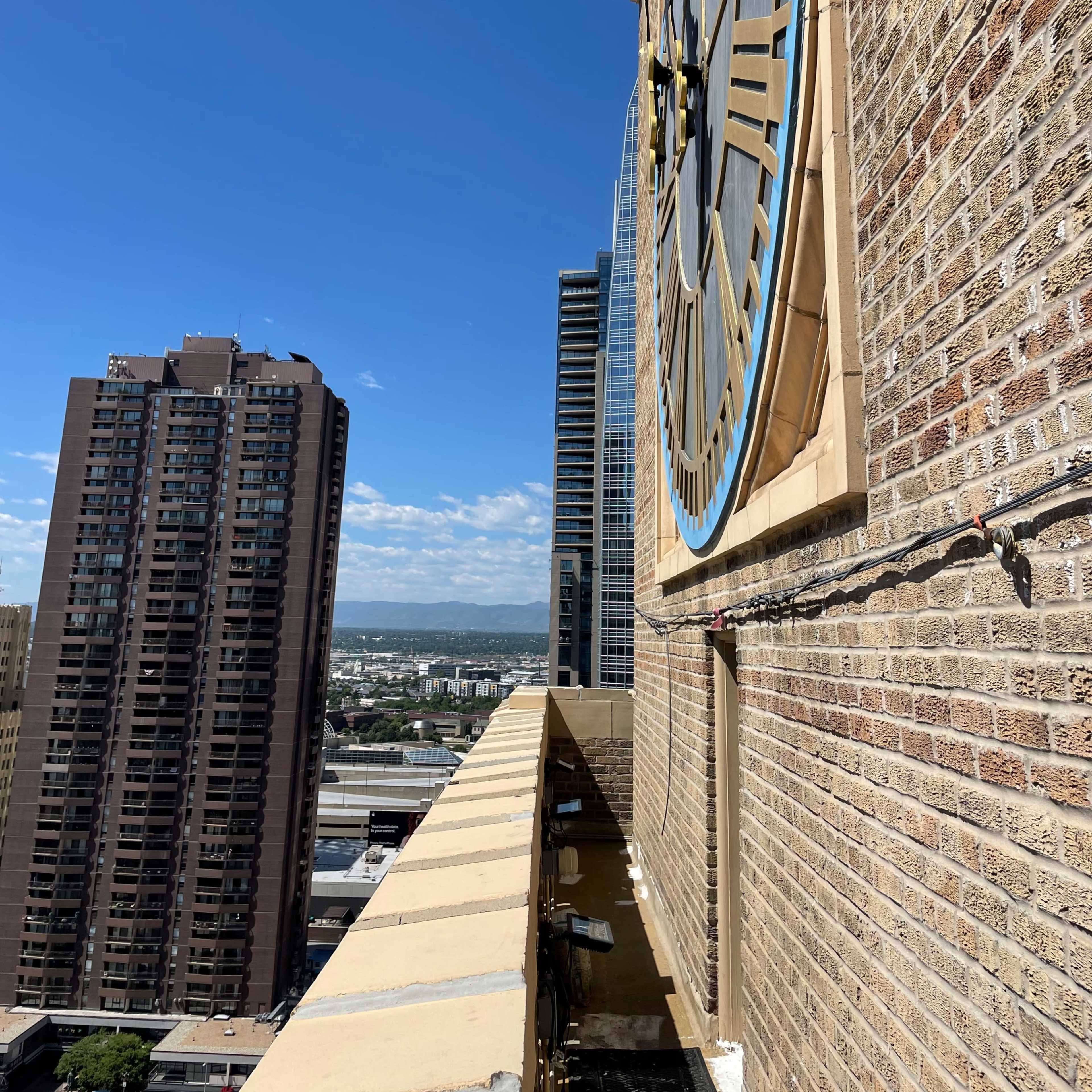 A close-up view of a large clock on a brick building, with a cityscape and blue sky in the background.