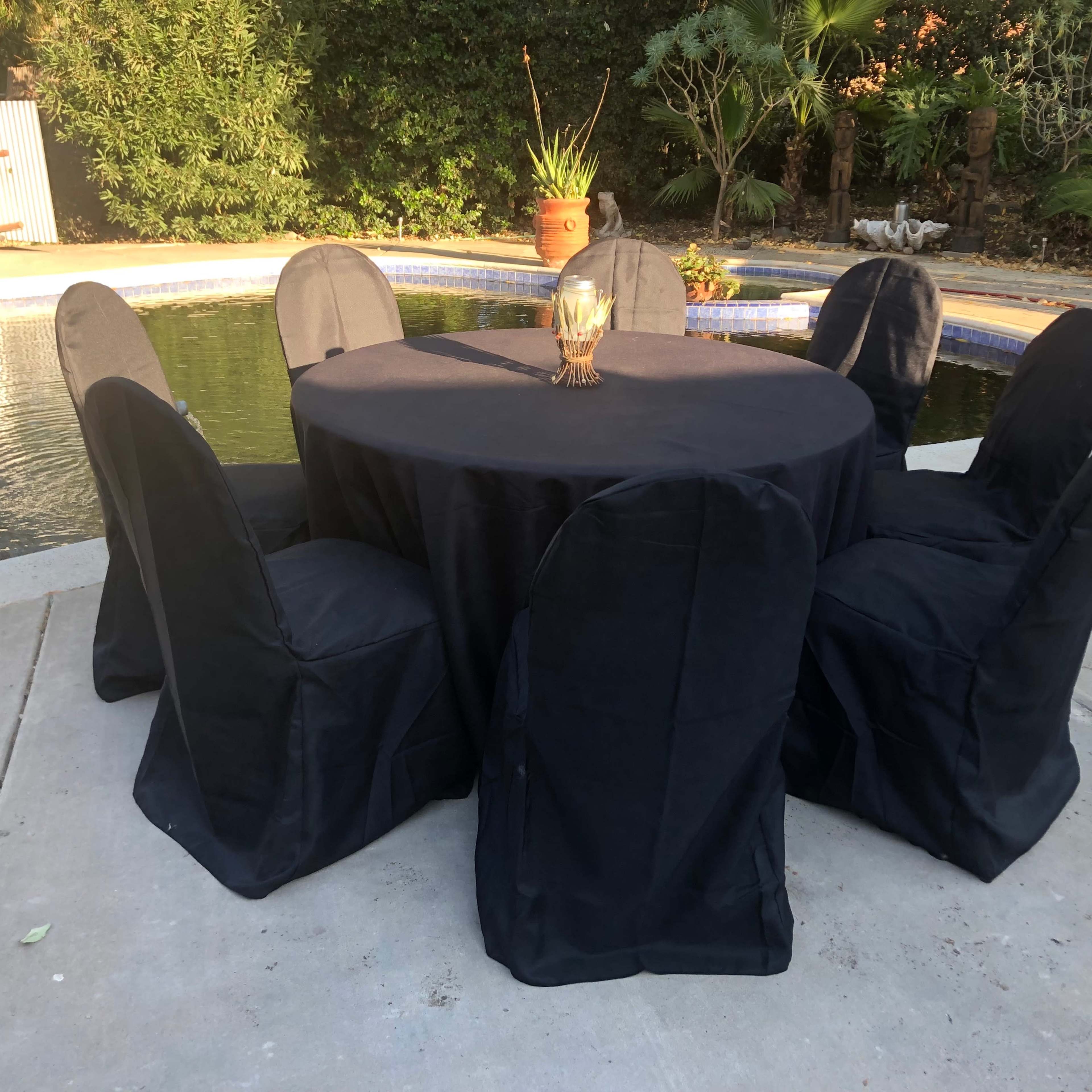A round table with a black tablecloth is set outdoors, surrounded by six black-covered chairs near a swimming pool and landscaped area.
