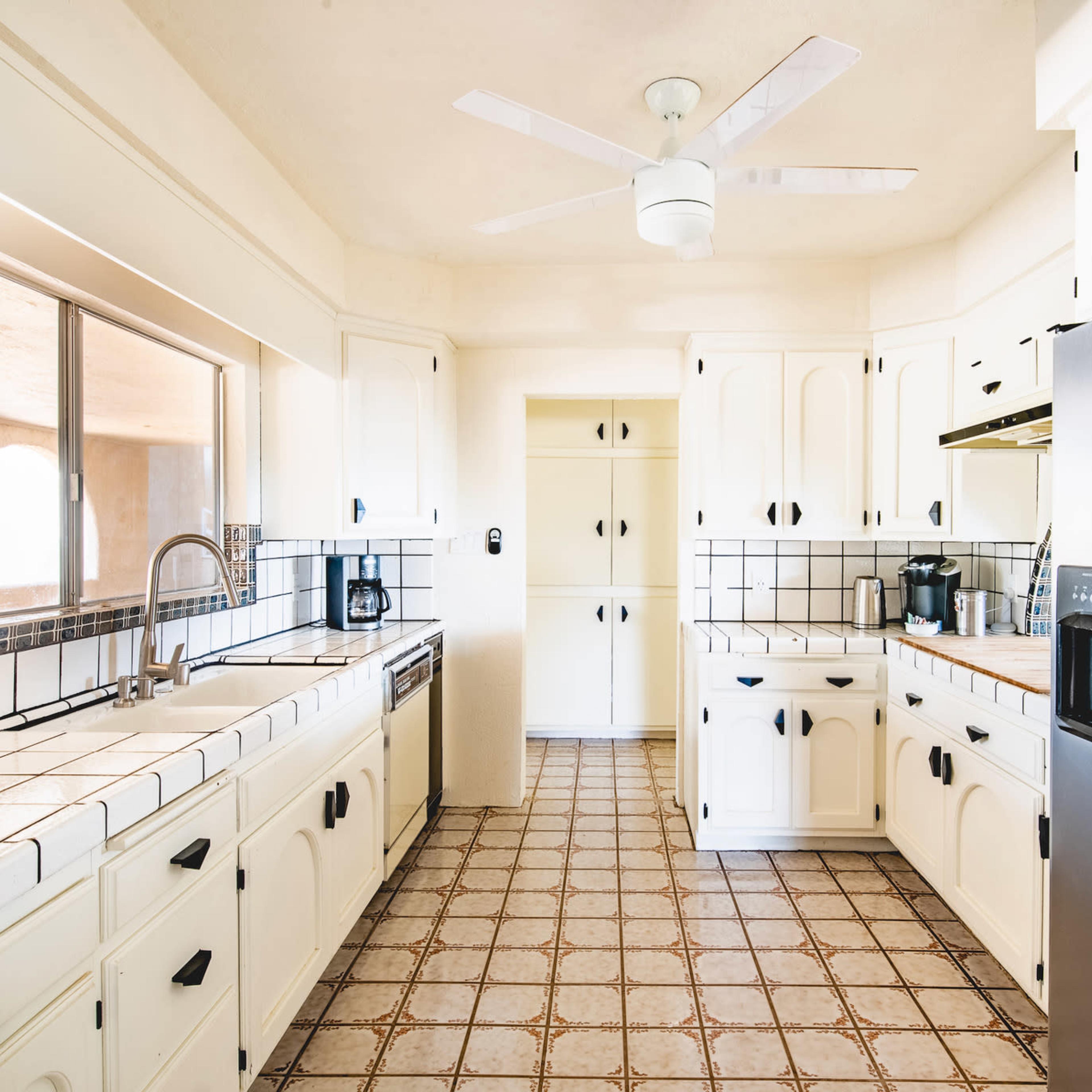 The kitchen features white cabinets, tiled countertops, and a stainless steel refrigerator, with a ceiling fan above and a window providing natural light.