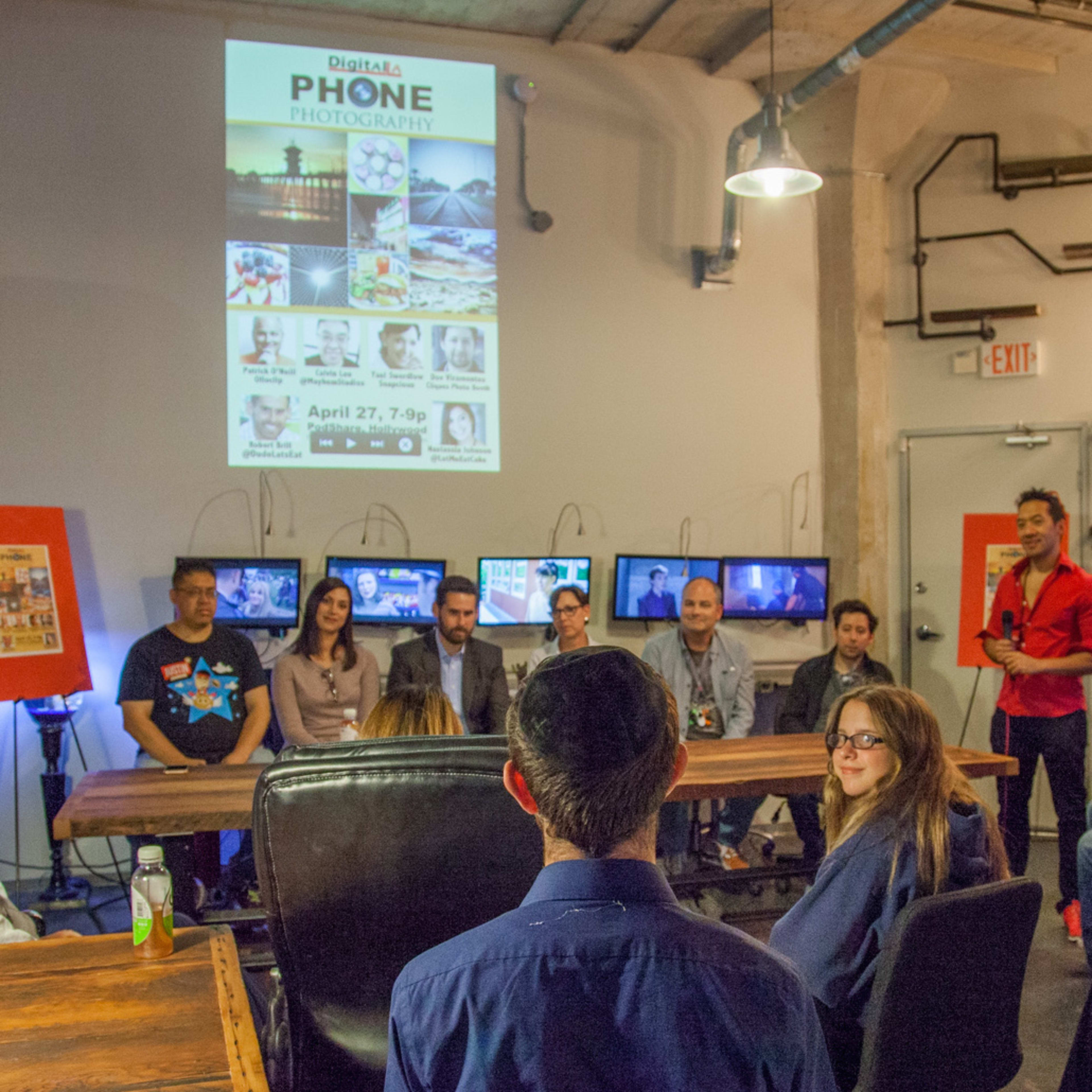 A group of people sits in a well-lit room with screens displaying photography, while a presenter stands in front of a large poster announcing a phone photography event.