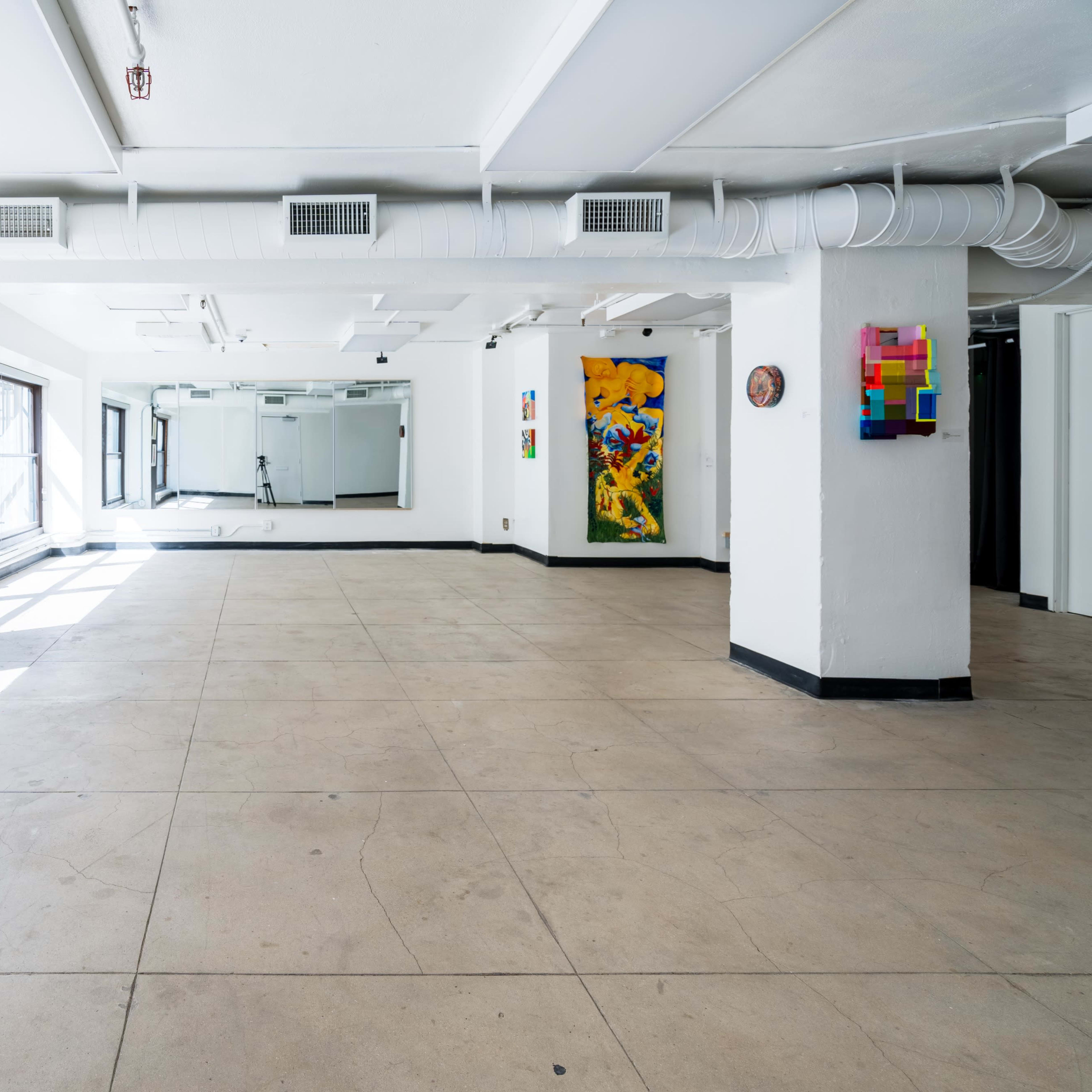 A spacious, empty gallery with large windows and white walls, featuring colorful artwork on two sides.