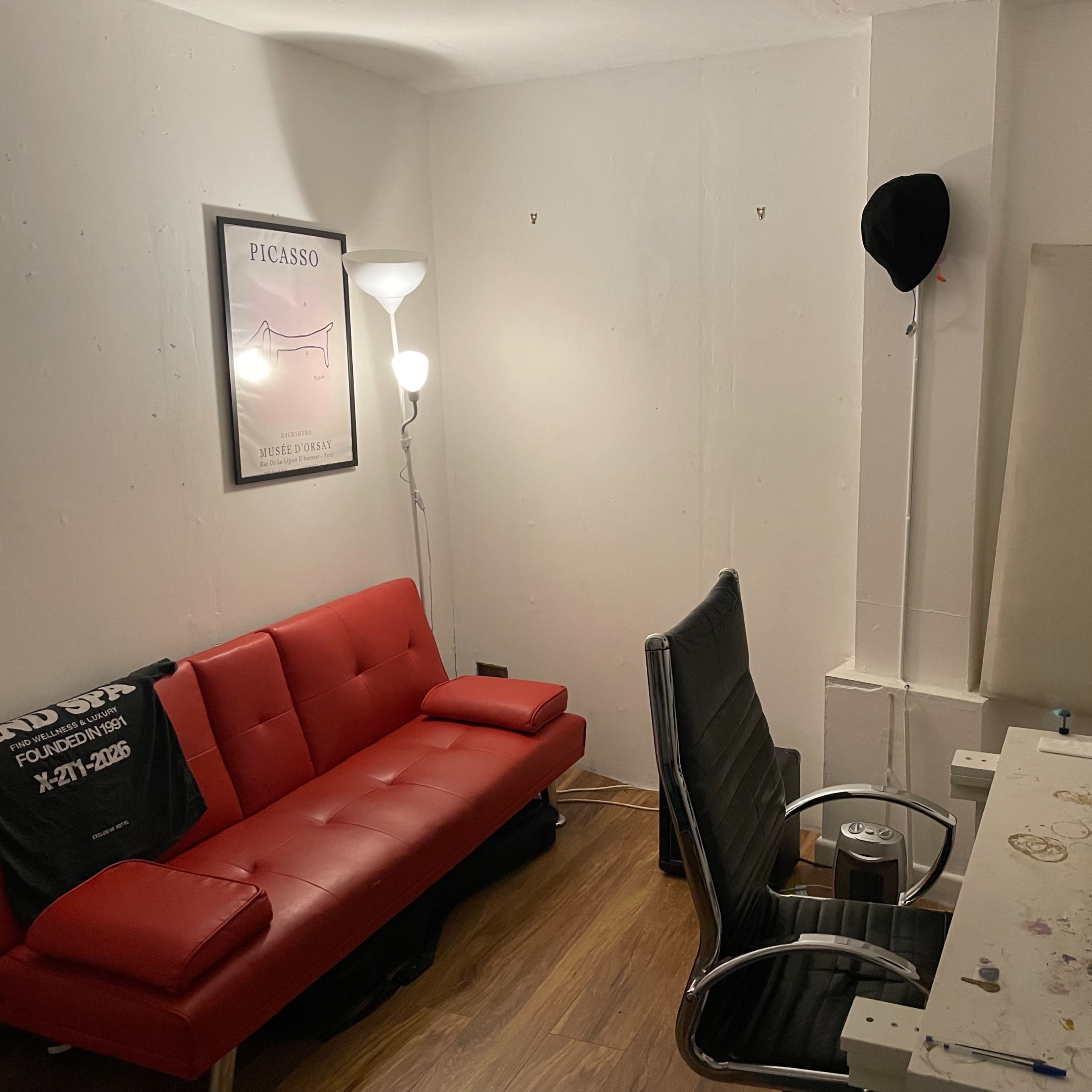 A small room with a red sofa, a black office chair, a keyboard on a desk, and a lamp, alongside a framed Picasso print on the wall.