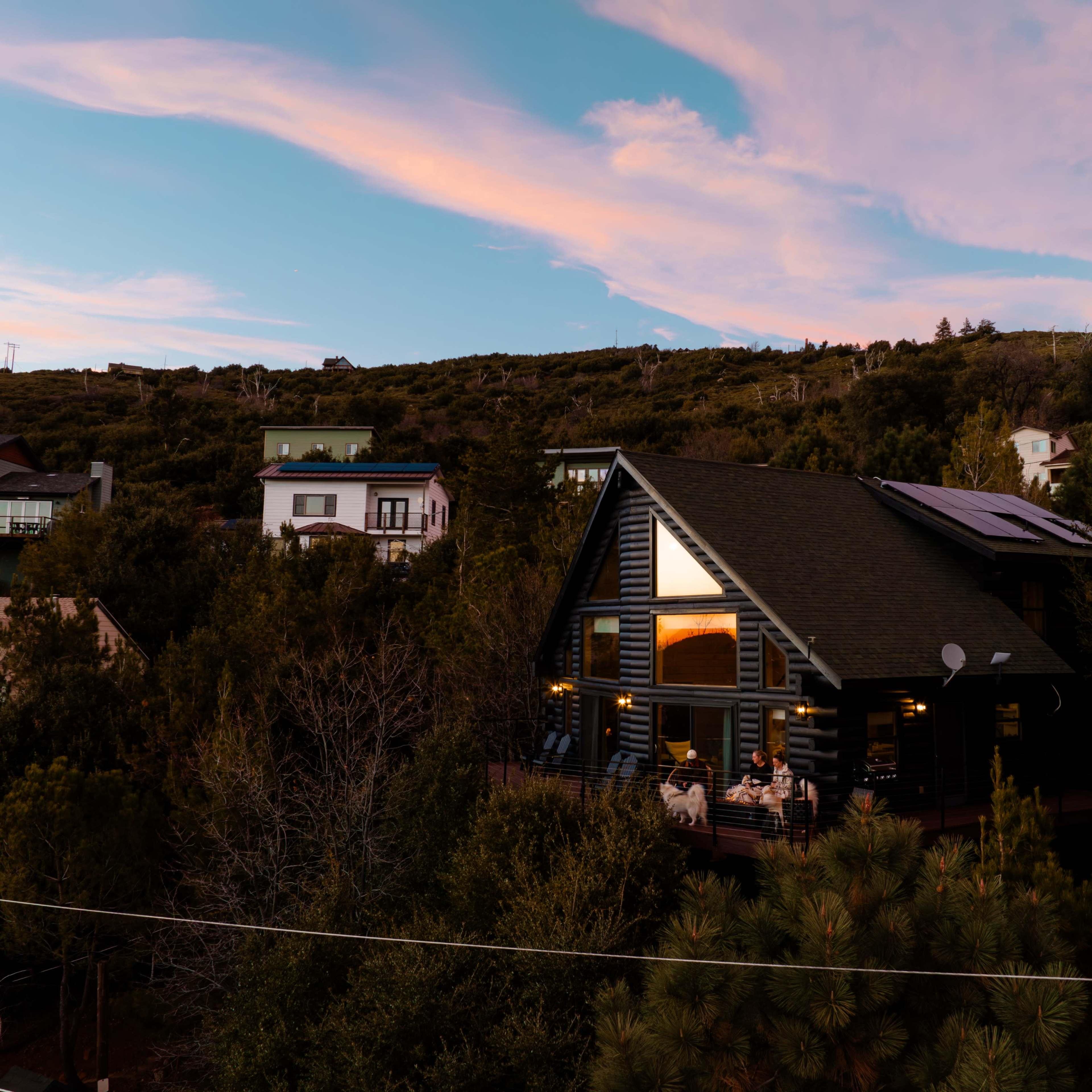 A dark wooden house with large windows sits among greenery, with people visible on the deck during sunset.