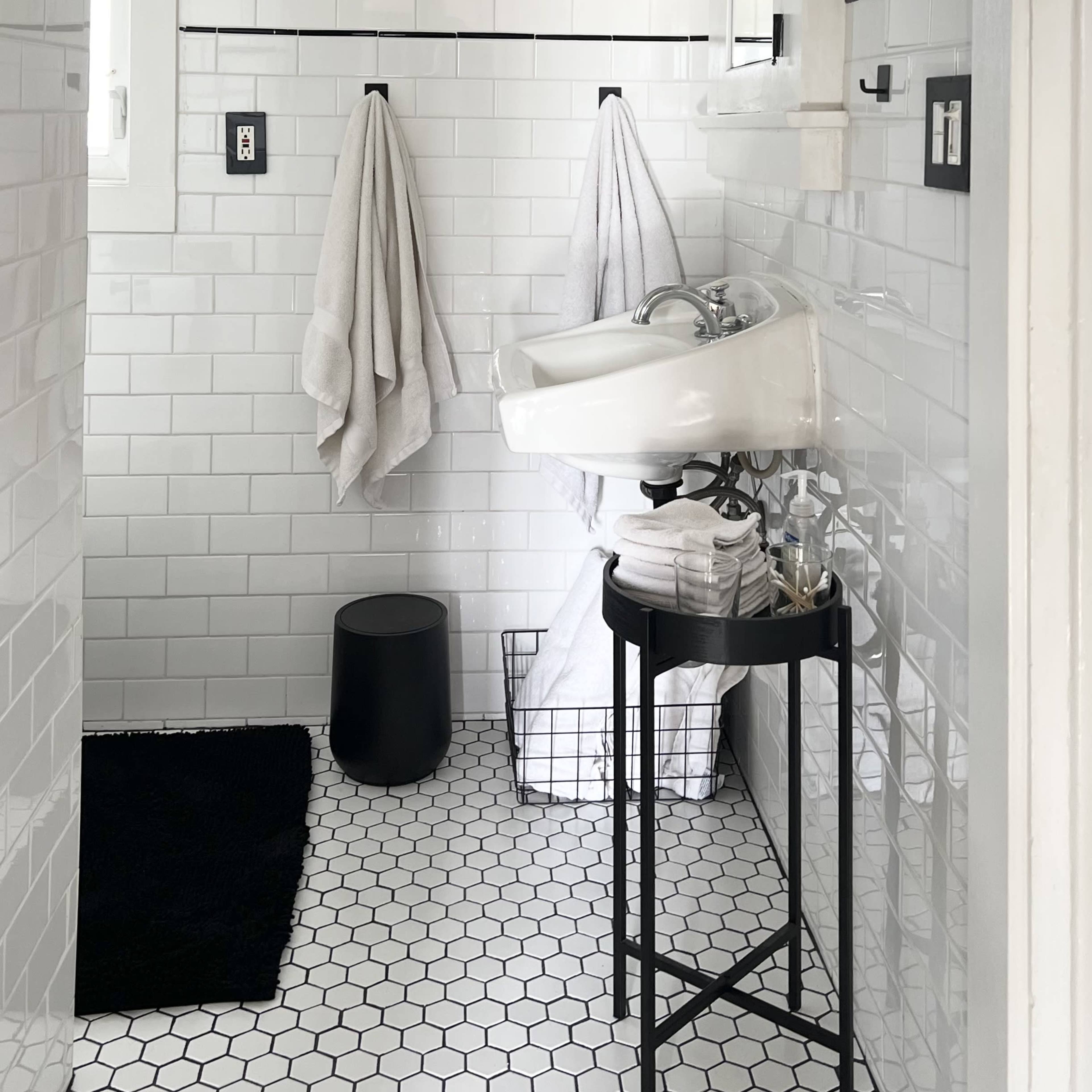 A modern bathroom featuring white tiled walls, a black and white photograph framed on the wall, a pedestal sink, and two towels hanging on hooks.