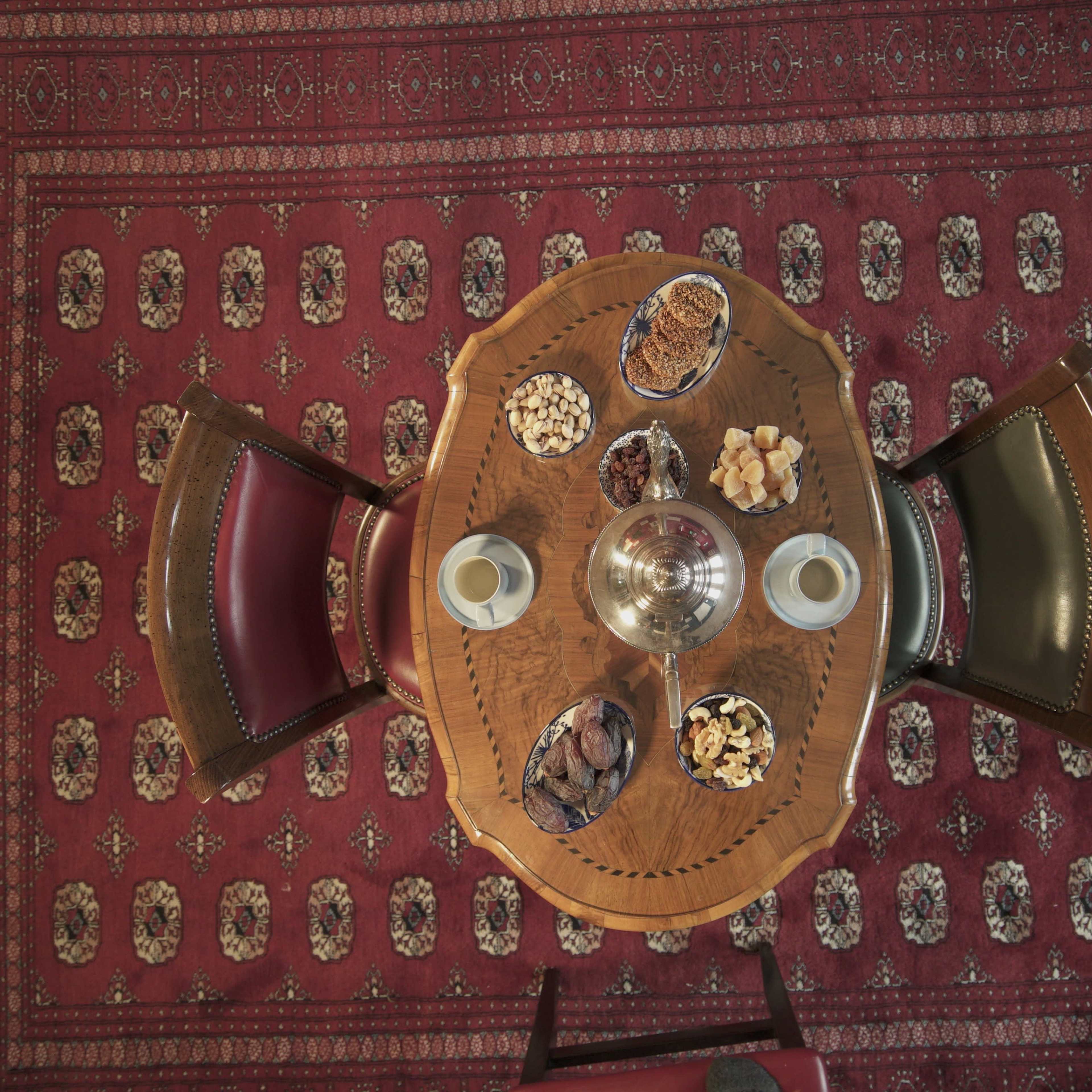 A round wooden table is set with various snacks and two cups, surrounded by two chairs, on a patterned red carpet.