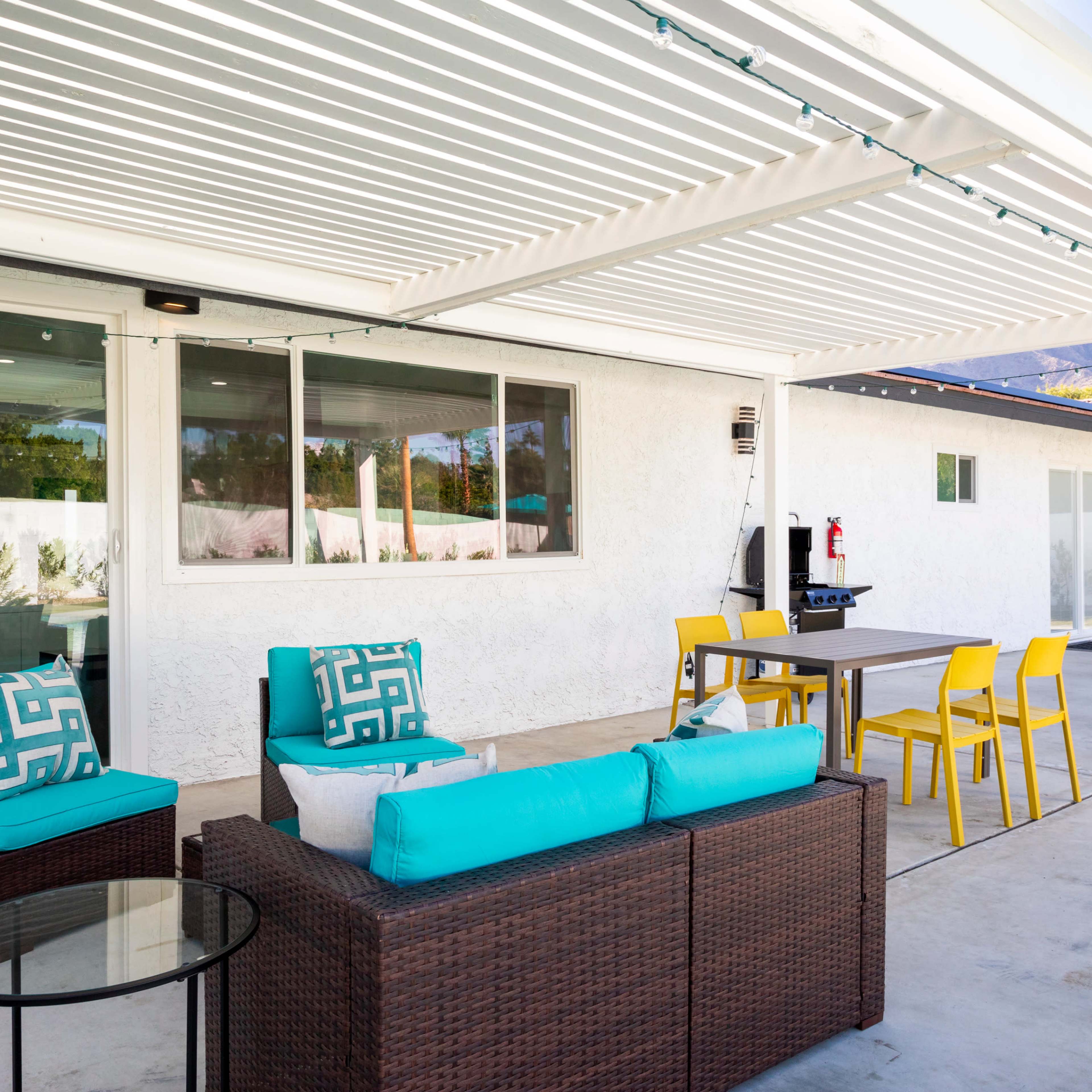 A patio area with a seating arrangement featuring turquoise cushions, a small table, a dining table with yellow chairs, and a sliding door leading into an interior space.