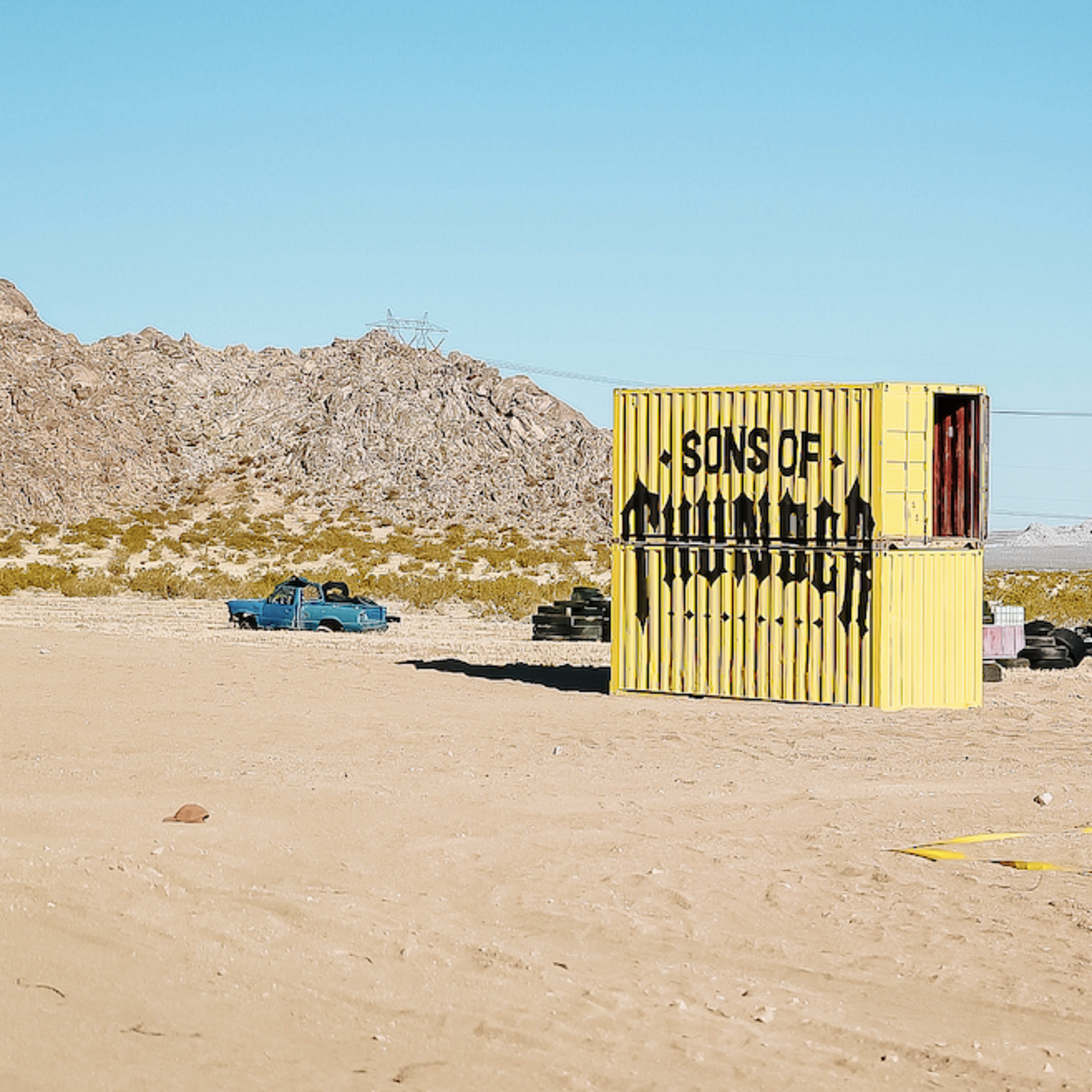 A yellow shipping container labeled "SONS OF" stands in a desert landscape surrounded by abandoned cars and rocky terrain.