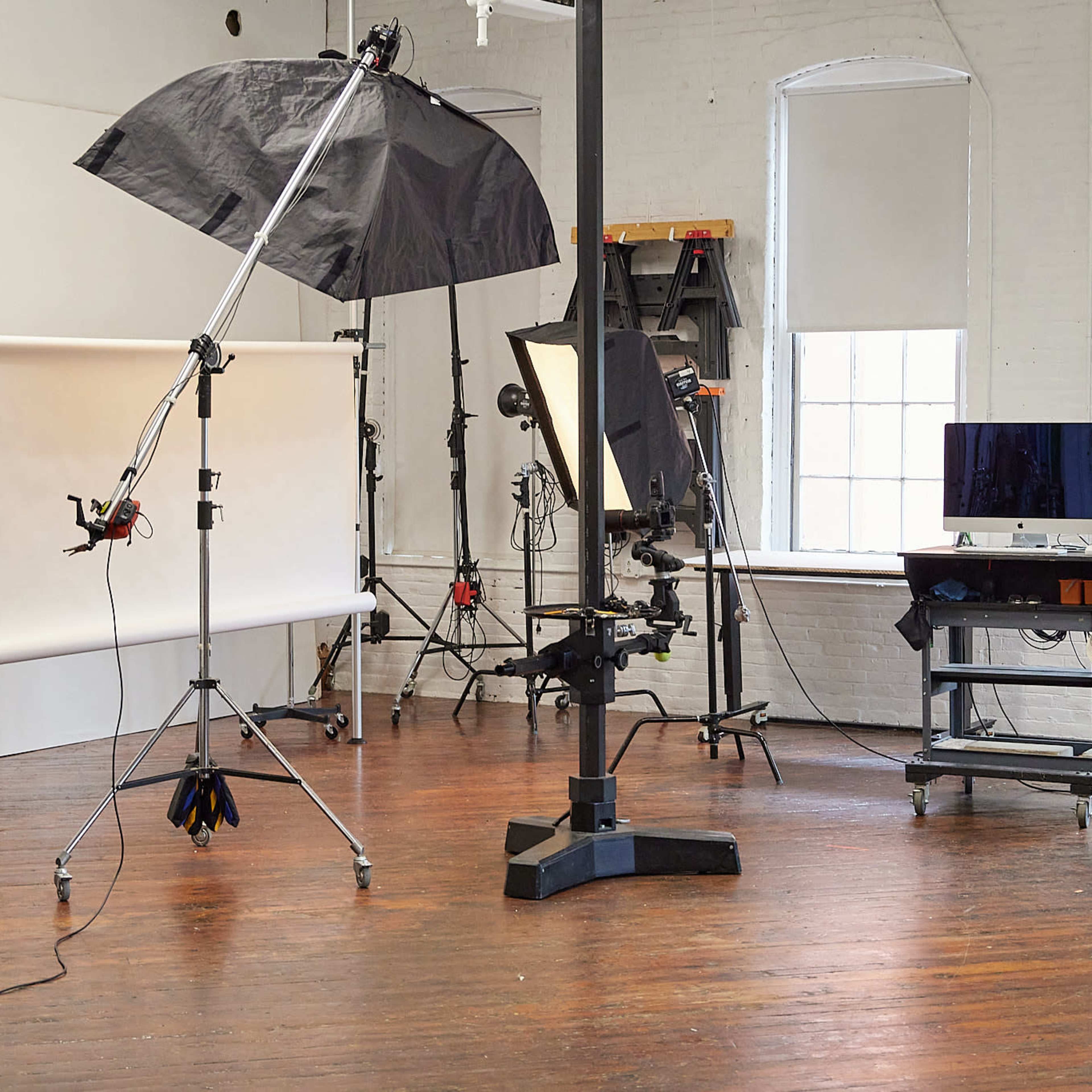 A photography studio setup with lighting equipment, a backdrop, and a small desk with a monitor.