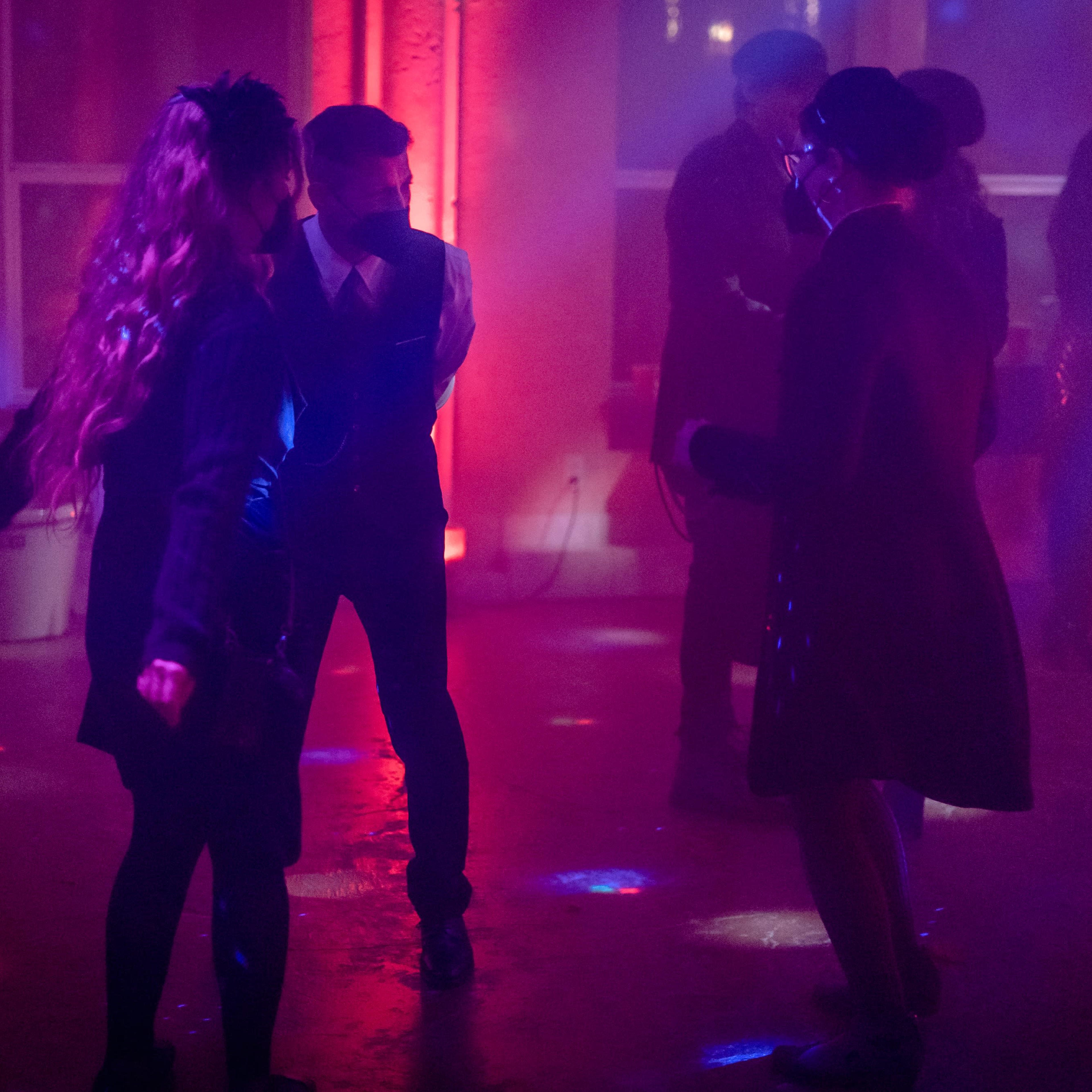 A group of people is dancing in a dimly lit venue with colorful lighting and a fog machine.