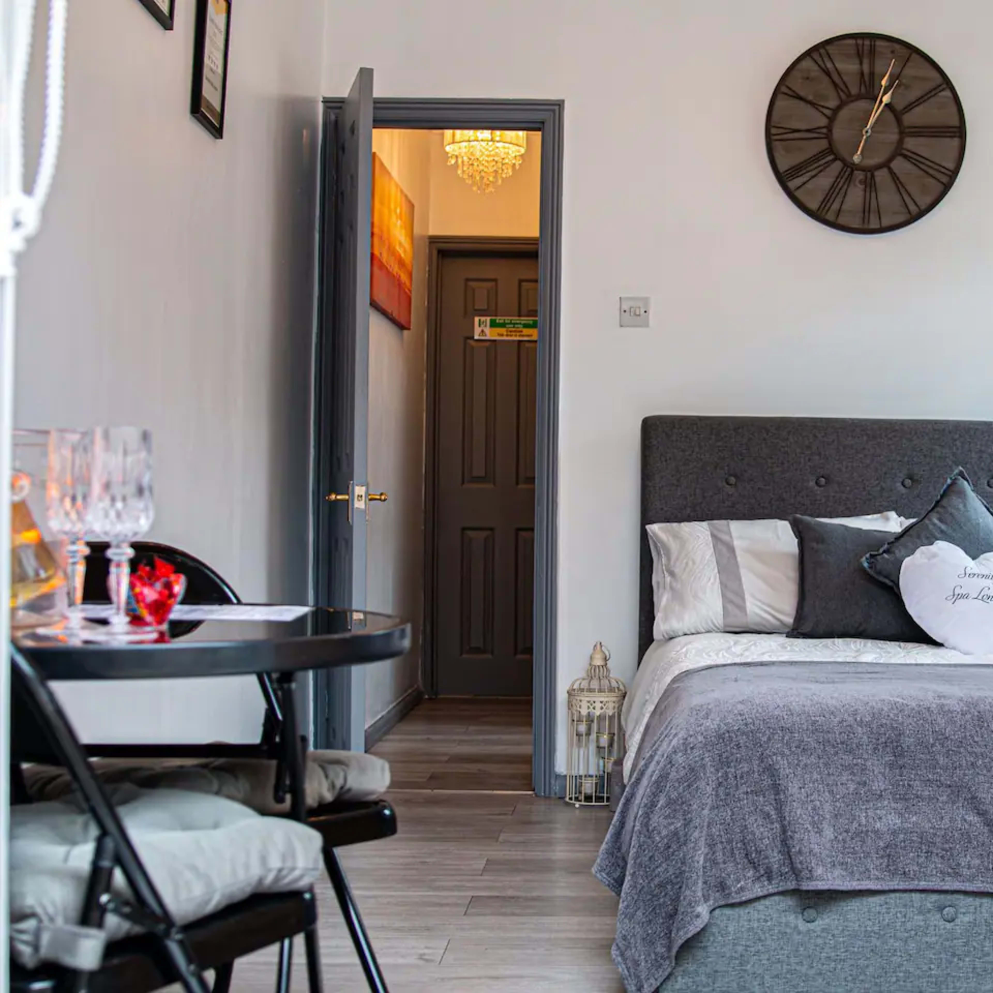 A compact room featuring a bed with gray bedding, a clock on the wall, a small dining table with chairs, and a doorway leading to another room.