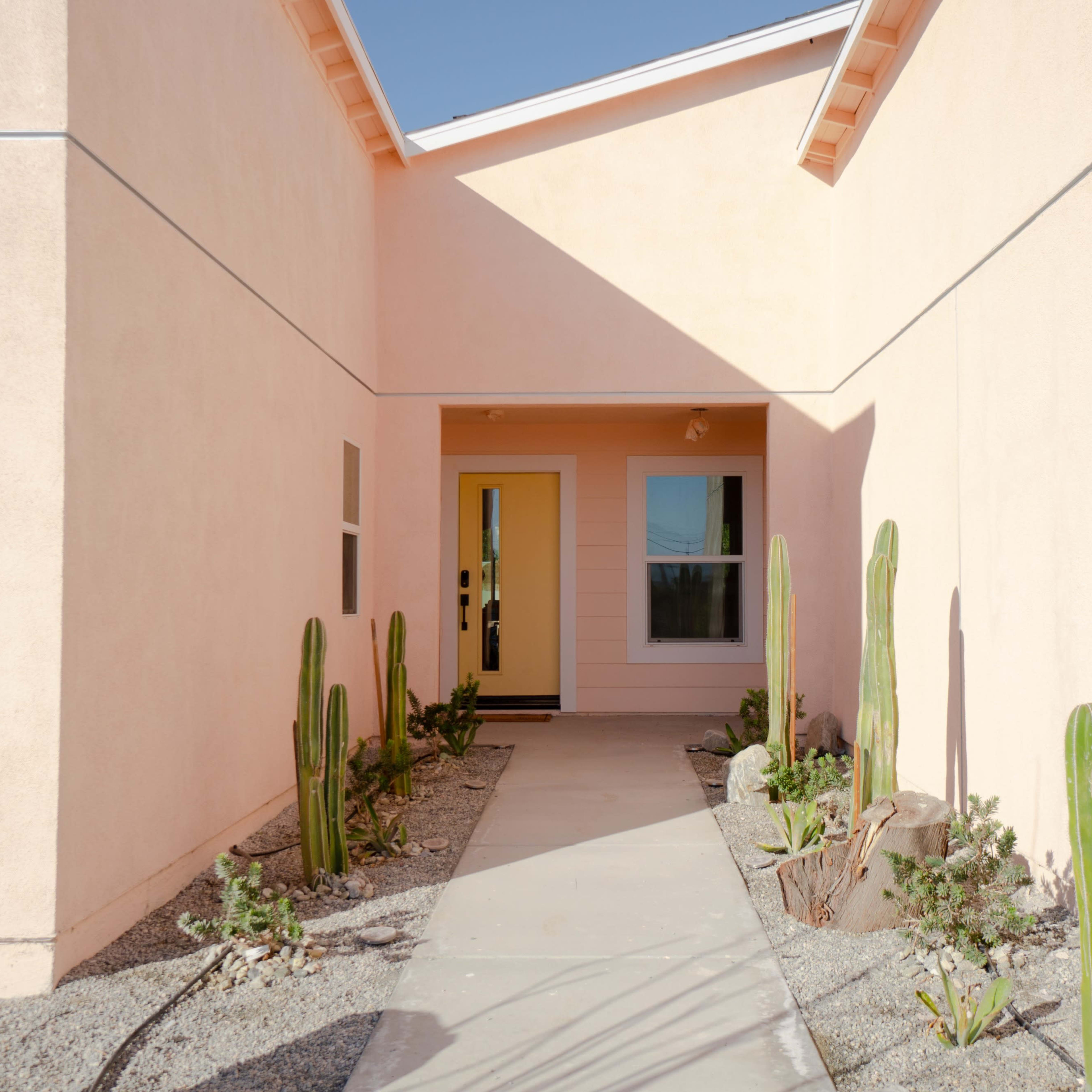 A narrow pathway leads to a yellow front door, flanked by tall cactus plants and bordered by gravel landscaping.