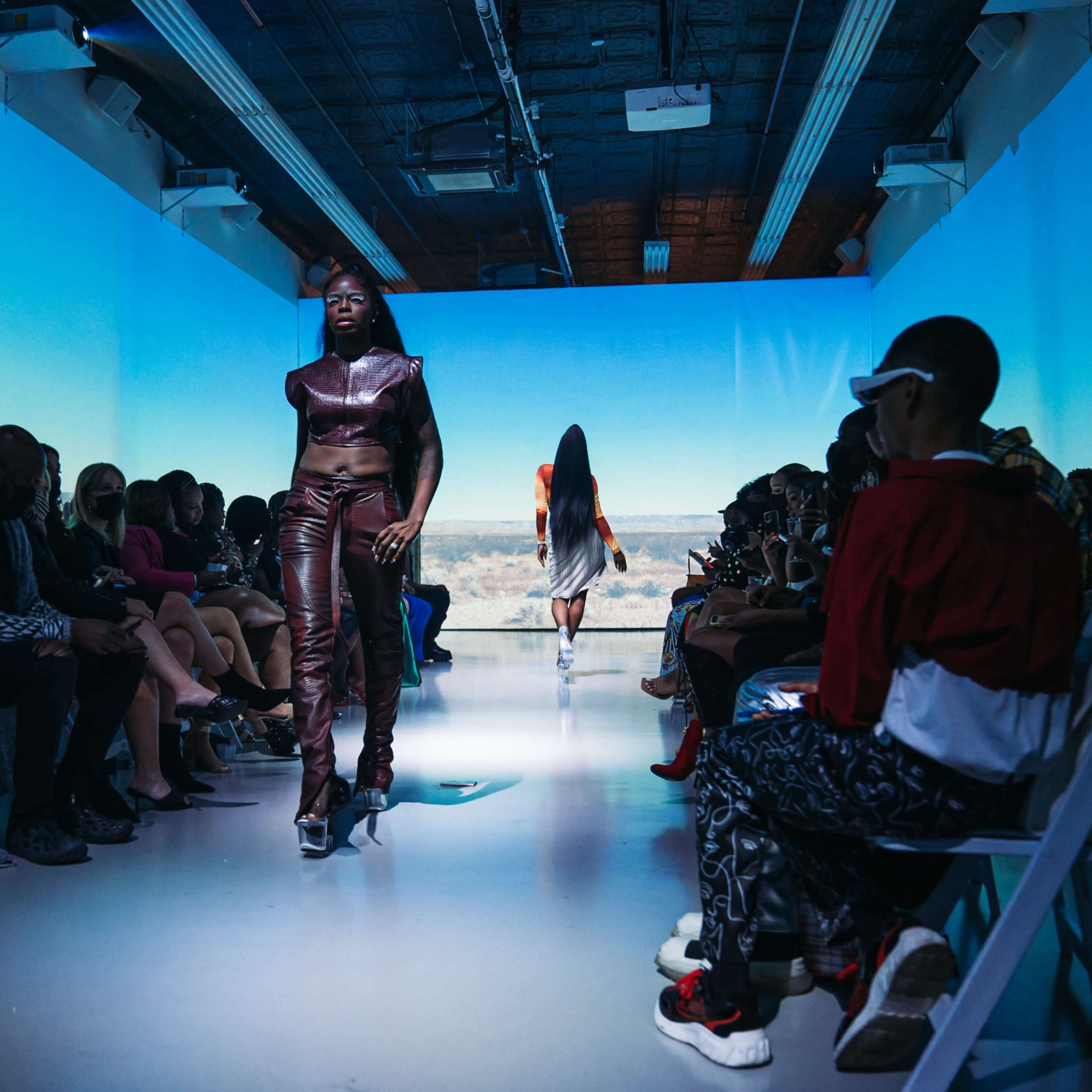 A fashion model walks down a runway surrounded by seated spectators as another model exits in the background.