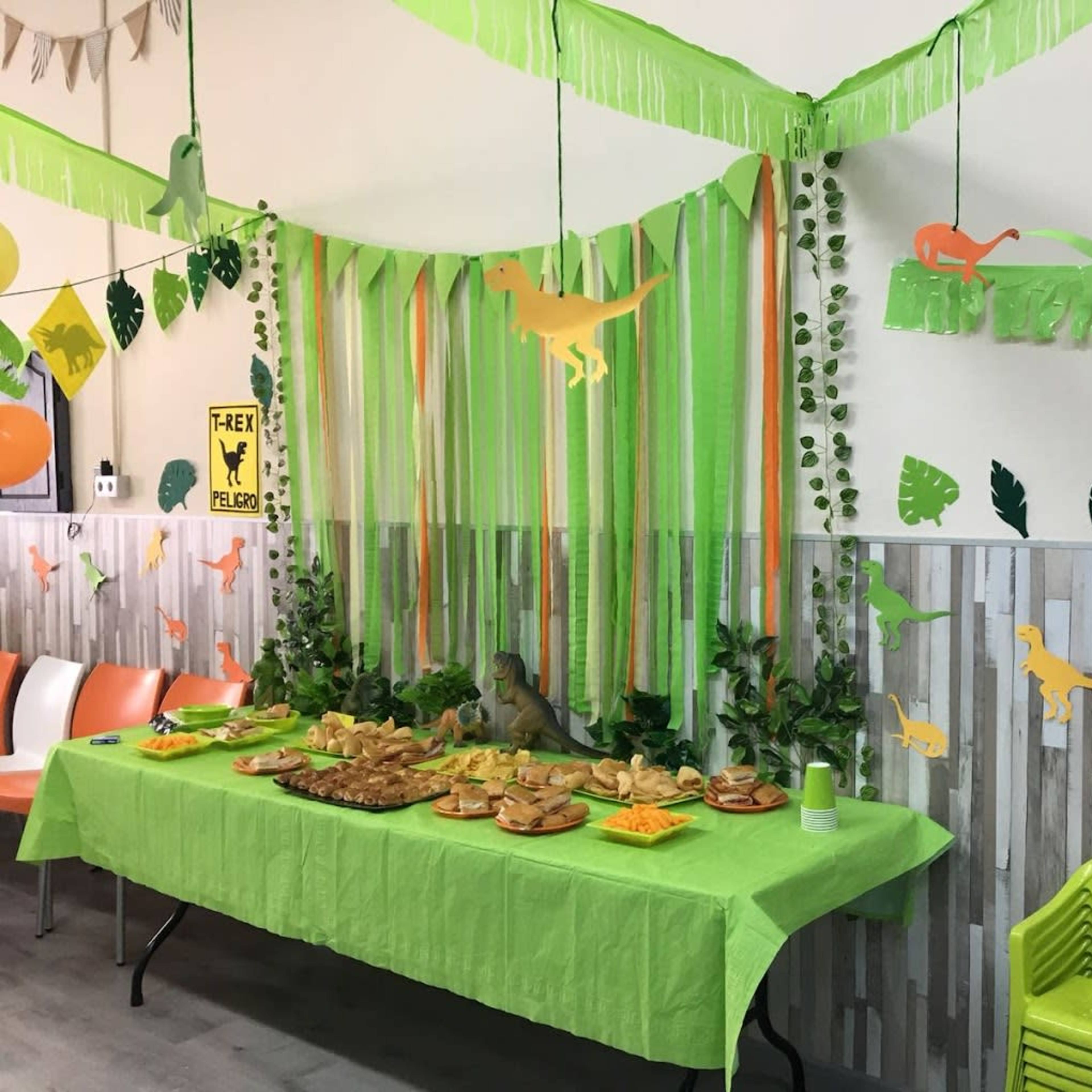 A themed party area features a green tablecloth with a spread of food, surrounded by dinosaur decorations and green streamers.