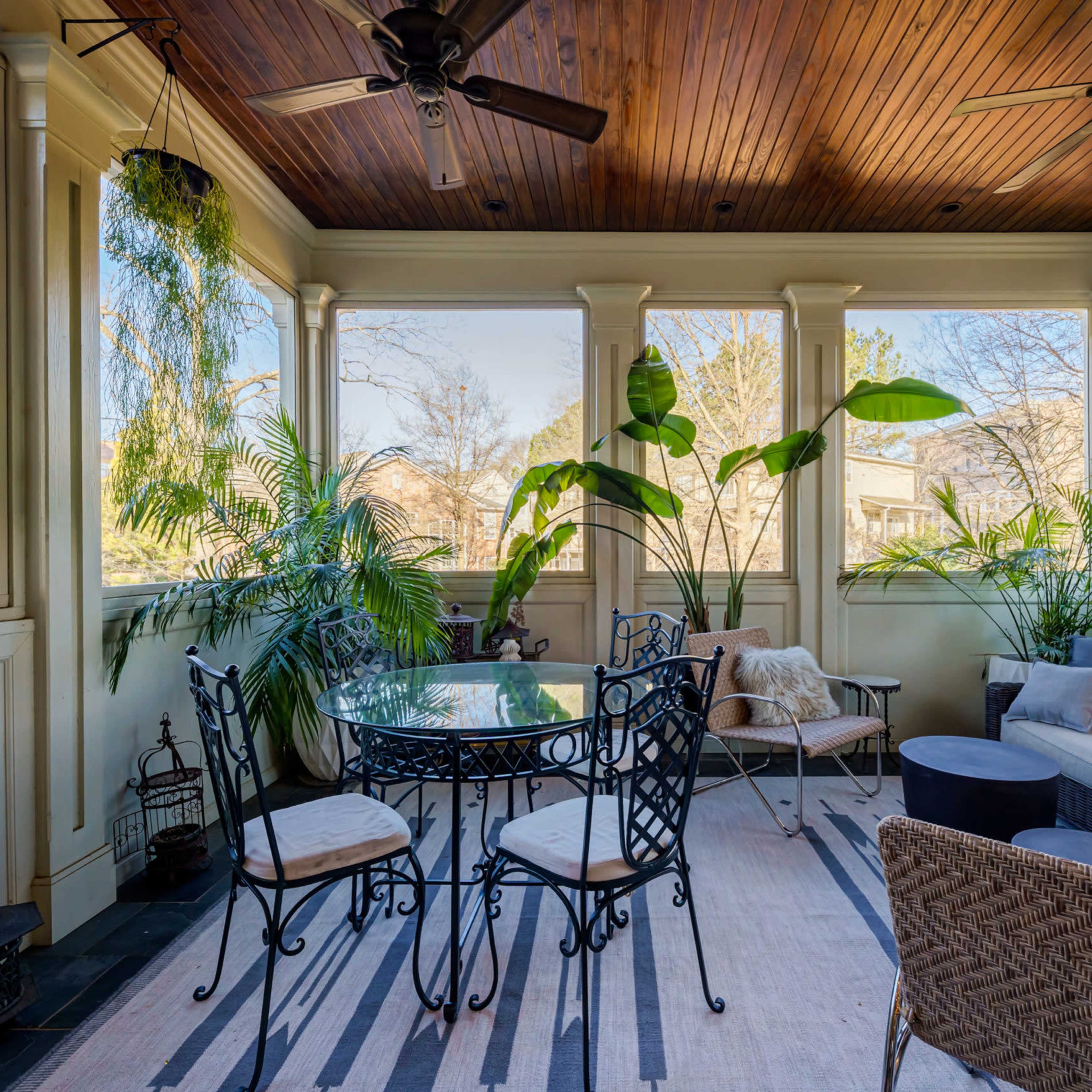A sunlit screened porch features a round glass table with metal chairs, a sectional sofa, and various indoor plants against wooden paneling and large windows.