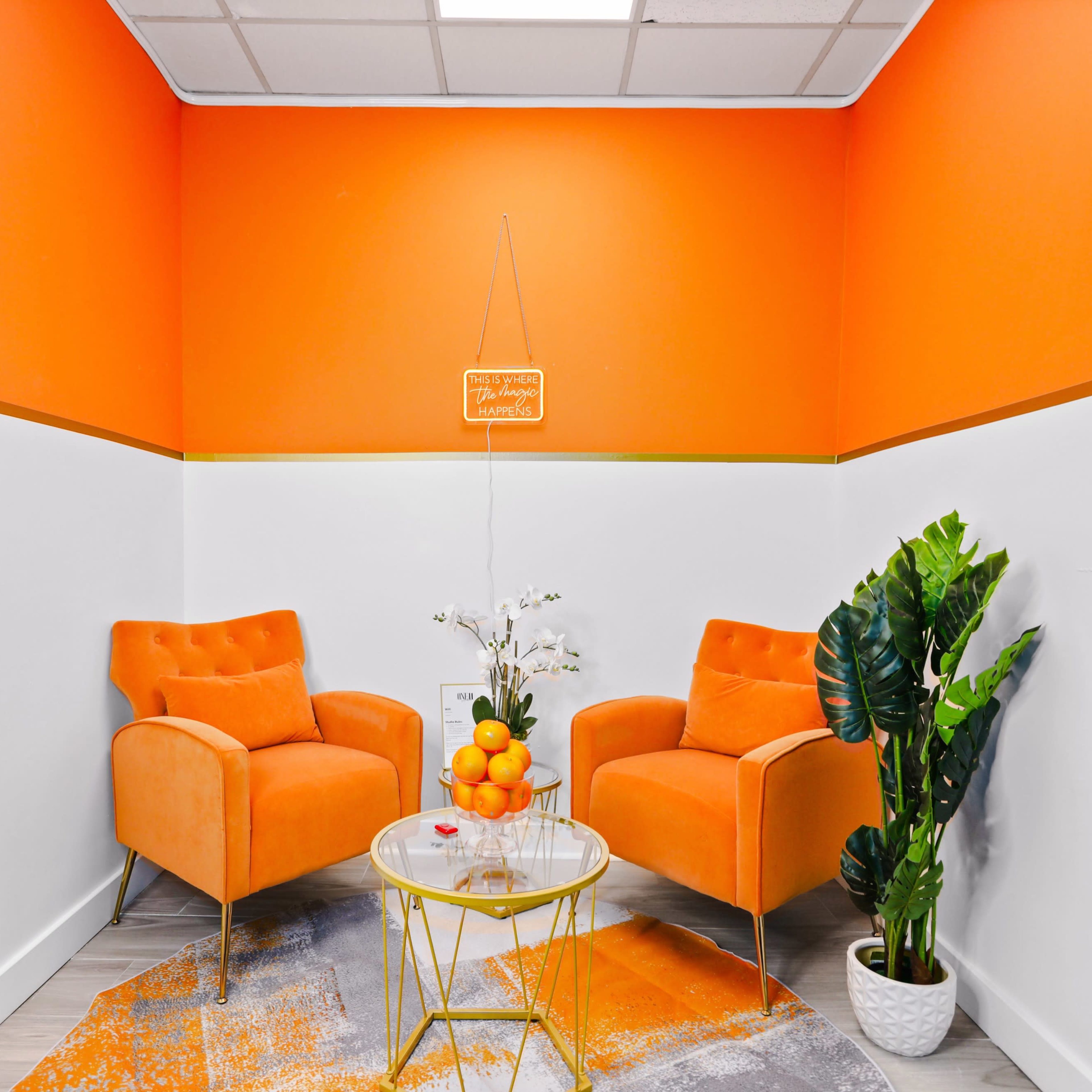 A sunlit corner with two orange chairs, a small glass table with a bowl of oranges, and a potted plant against a bright orange wall.