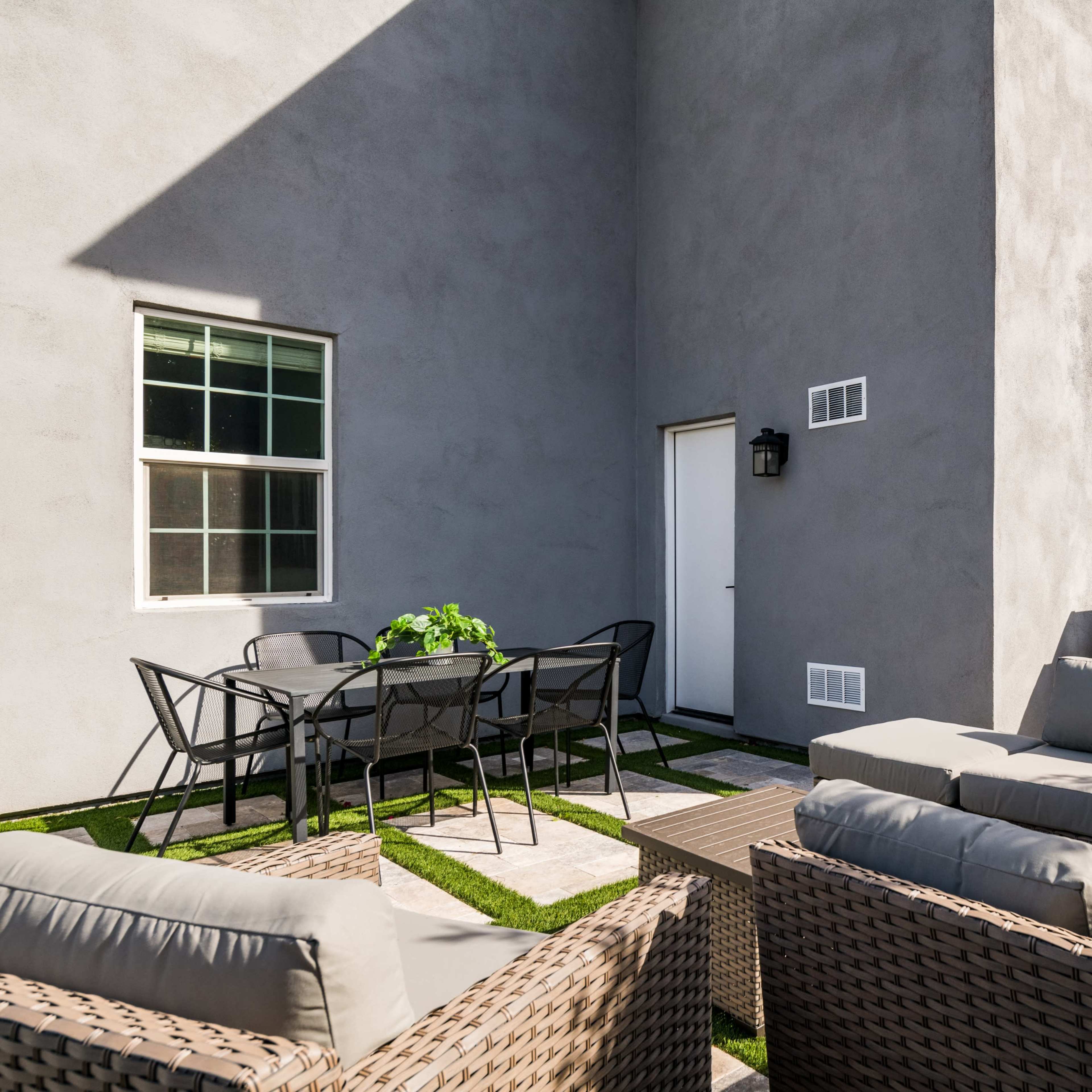 A patio area with wicker furniture, a dining table, and a grill next to a gray wall of a house.