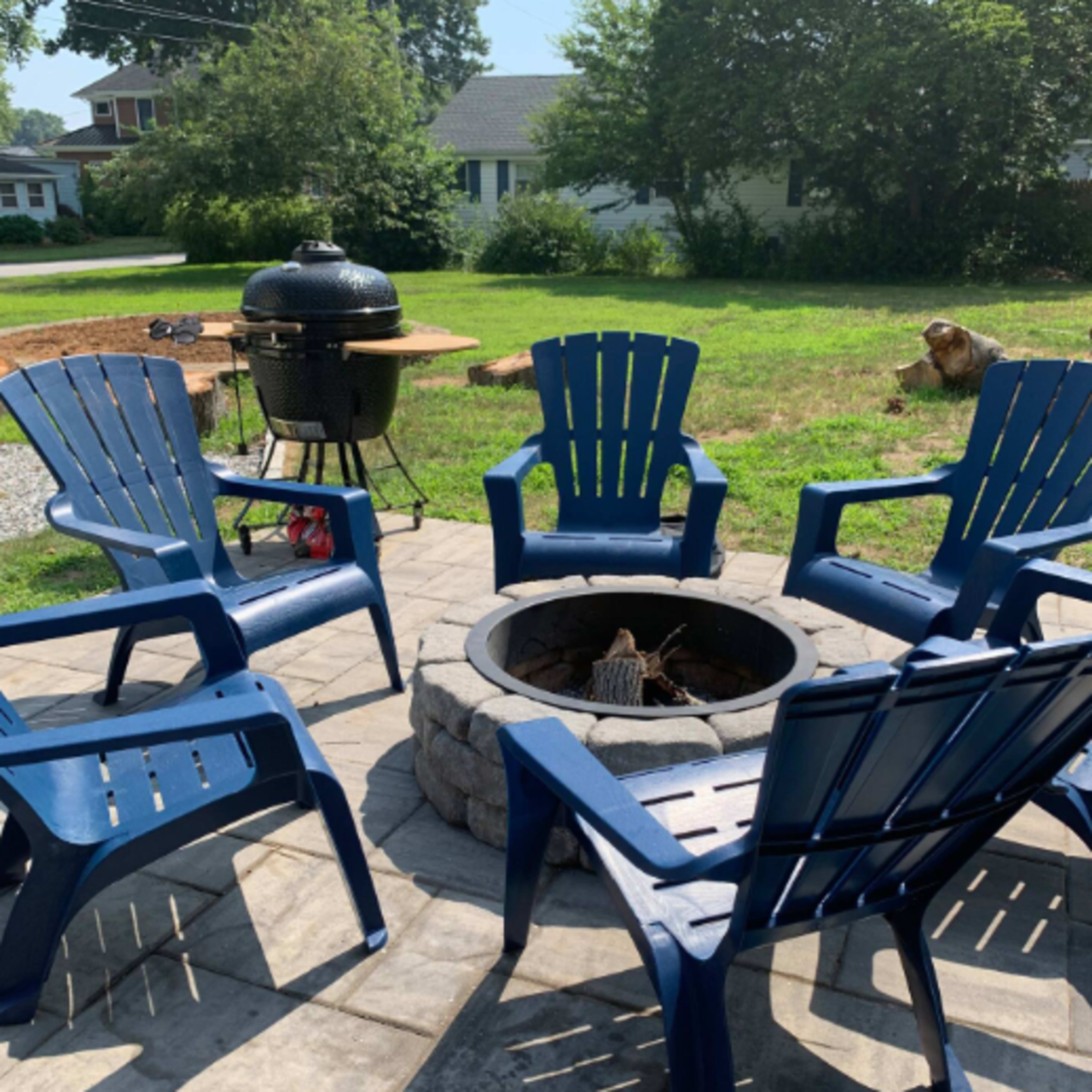 A stone fire pit is surrounded by blue adirondack chairs in a backyard with a grill and a dog in the background.