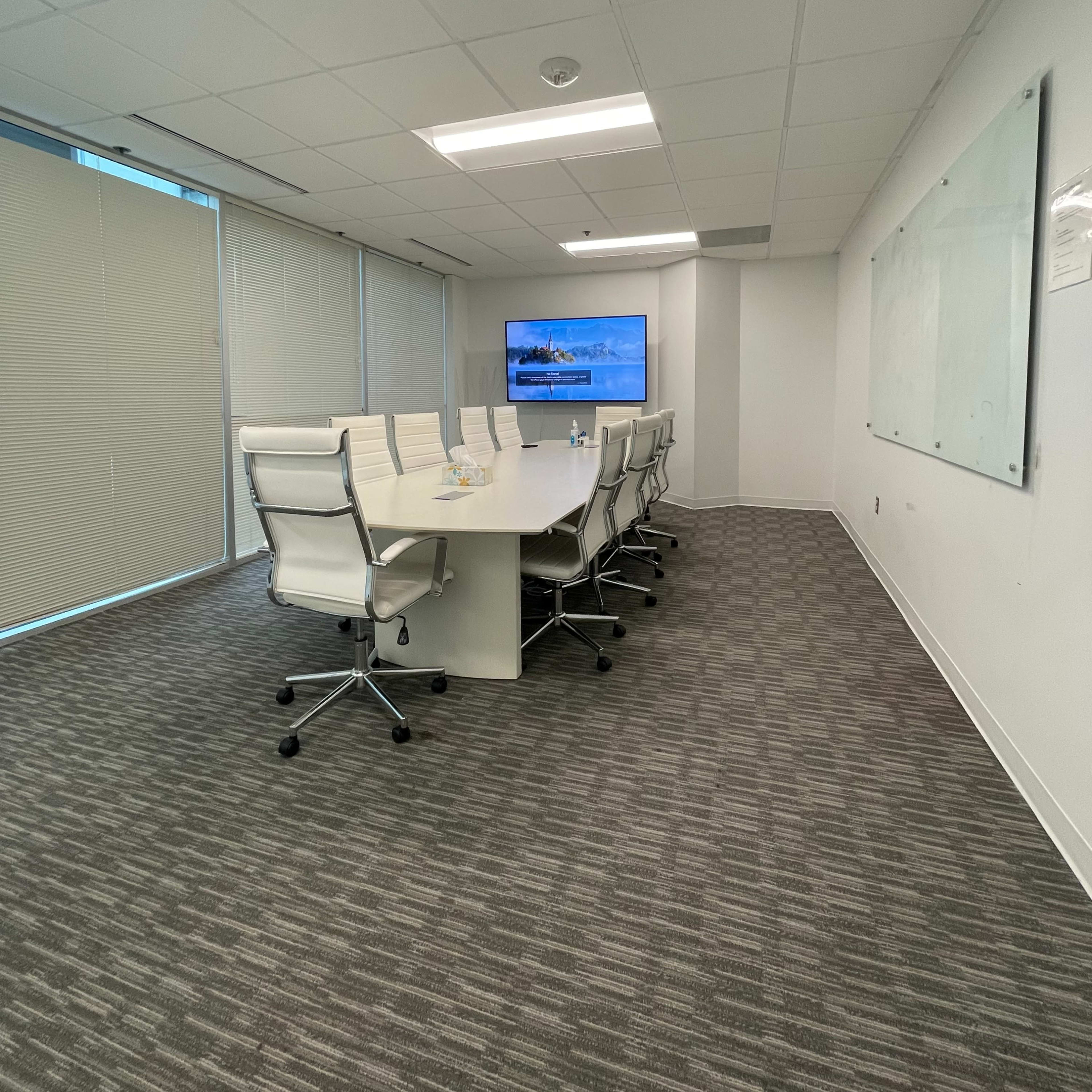 A conference room features a long white table with eight chairs, a large screen on the wall, and blinds covering the windows.