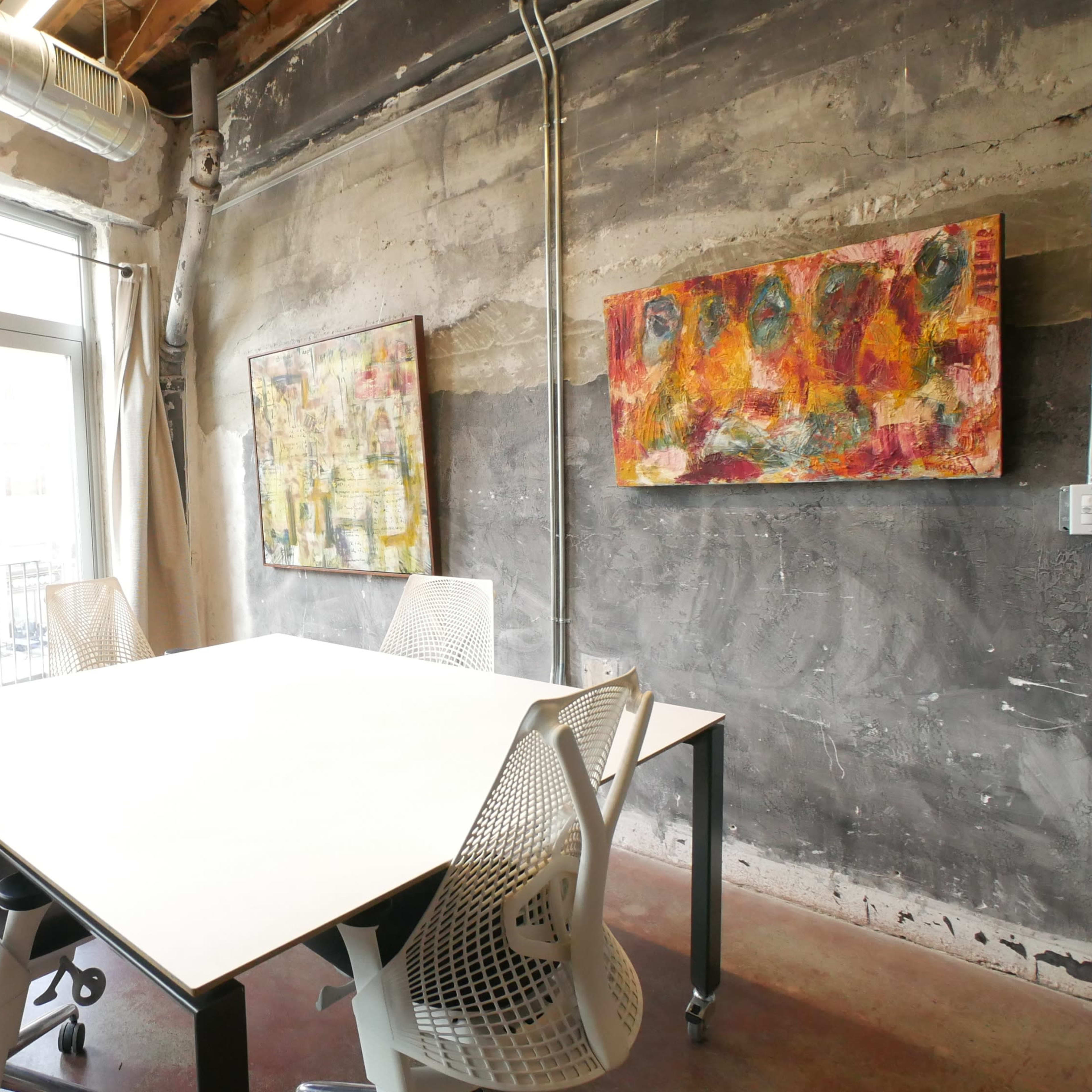 A modern meeting room with a large white table and four chairs, featuring exposed brick walls and two colorful paintings.