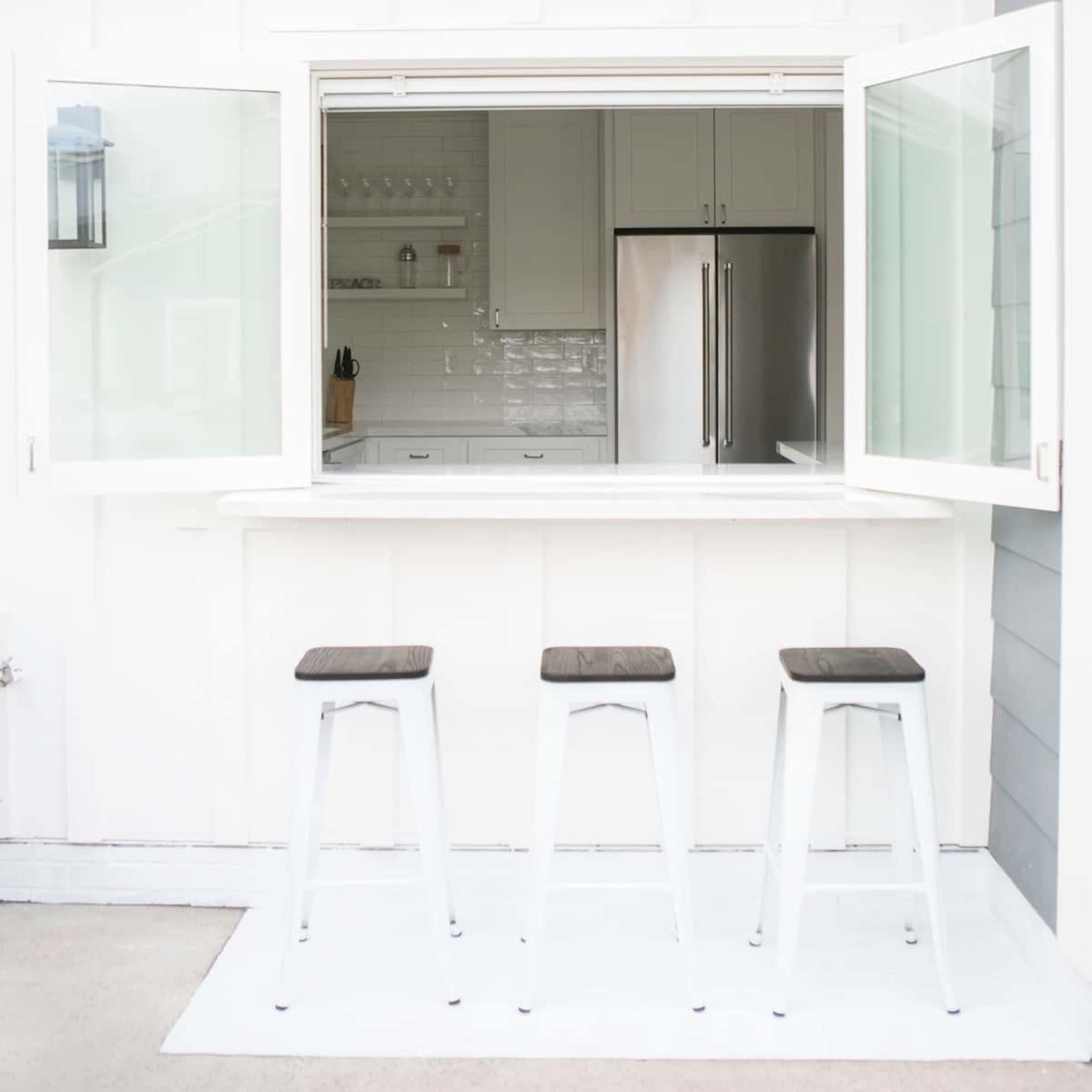 A white outdoor bar area features three stools in front of an open window, revealing a modern kitchen with stainless steel appliances.