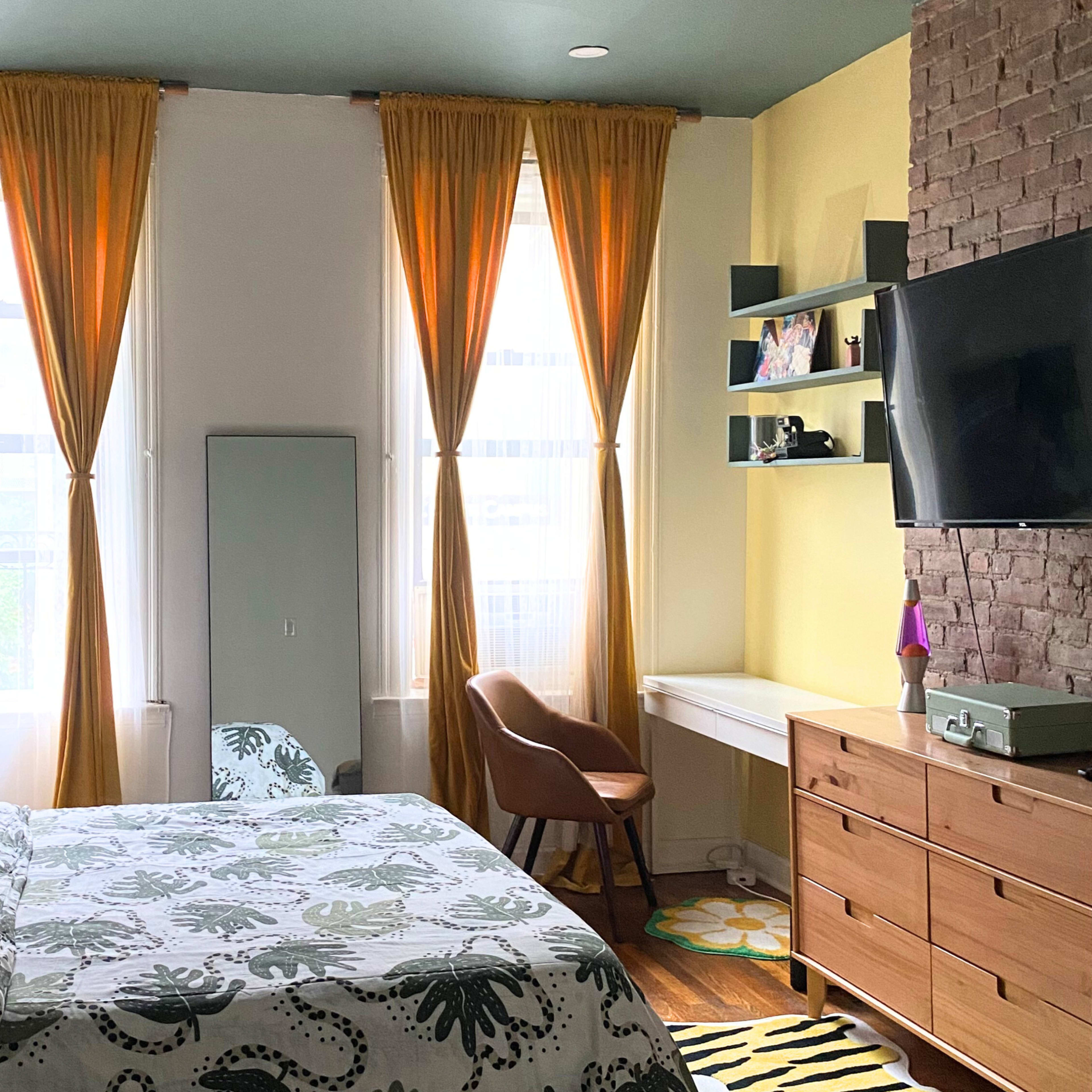 A bedroom with a bed, a desk, a chair, and large windows covered with yellow curtains, all set against a backdrop of light yellow and brick walls.