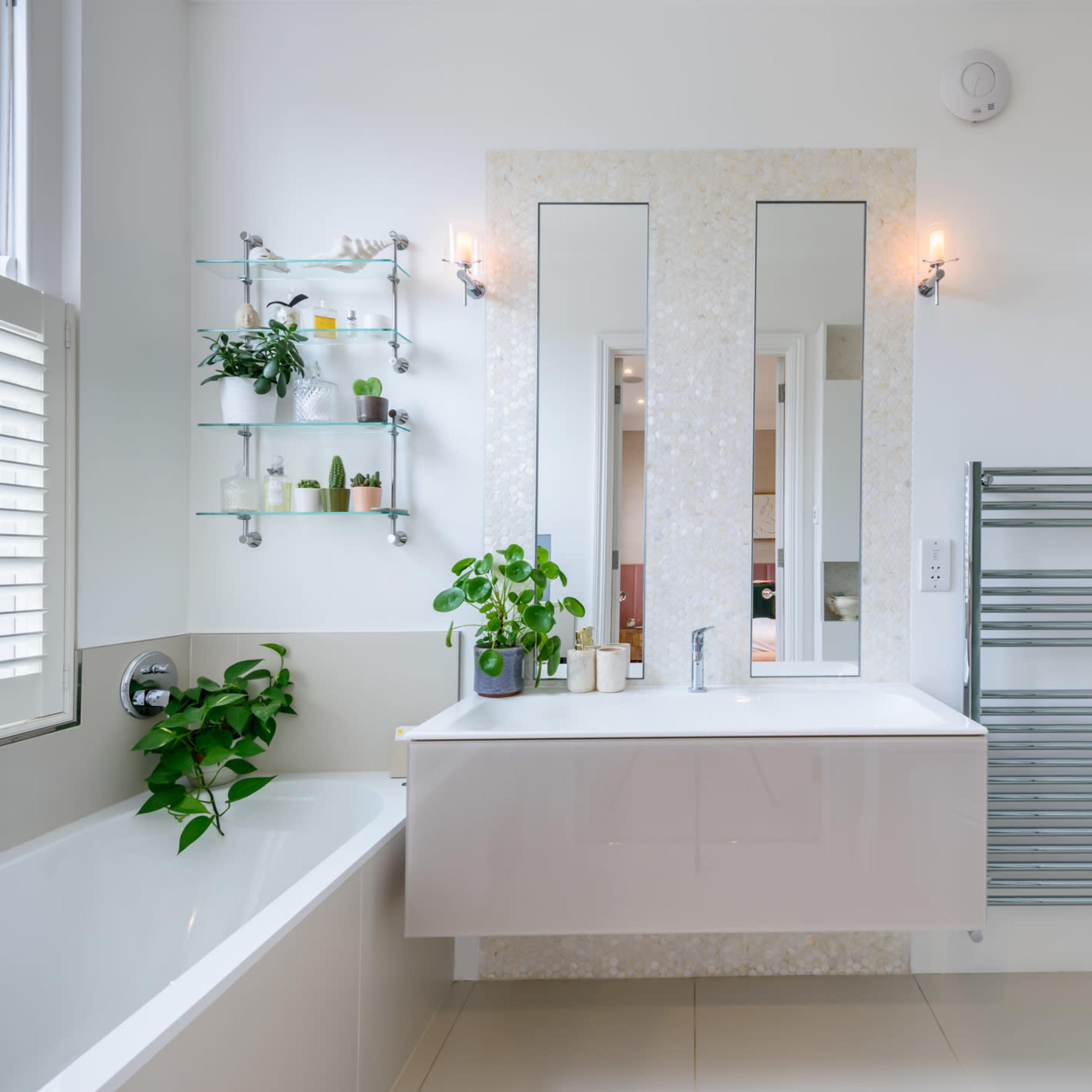 A modern bathroom featuring a bathtub, a wall-mounted sink with a mirror above, a glass shower enclosure, and plants on a shelf.