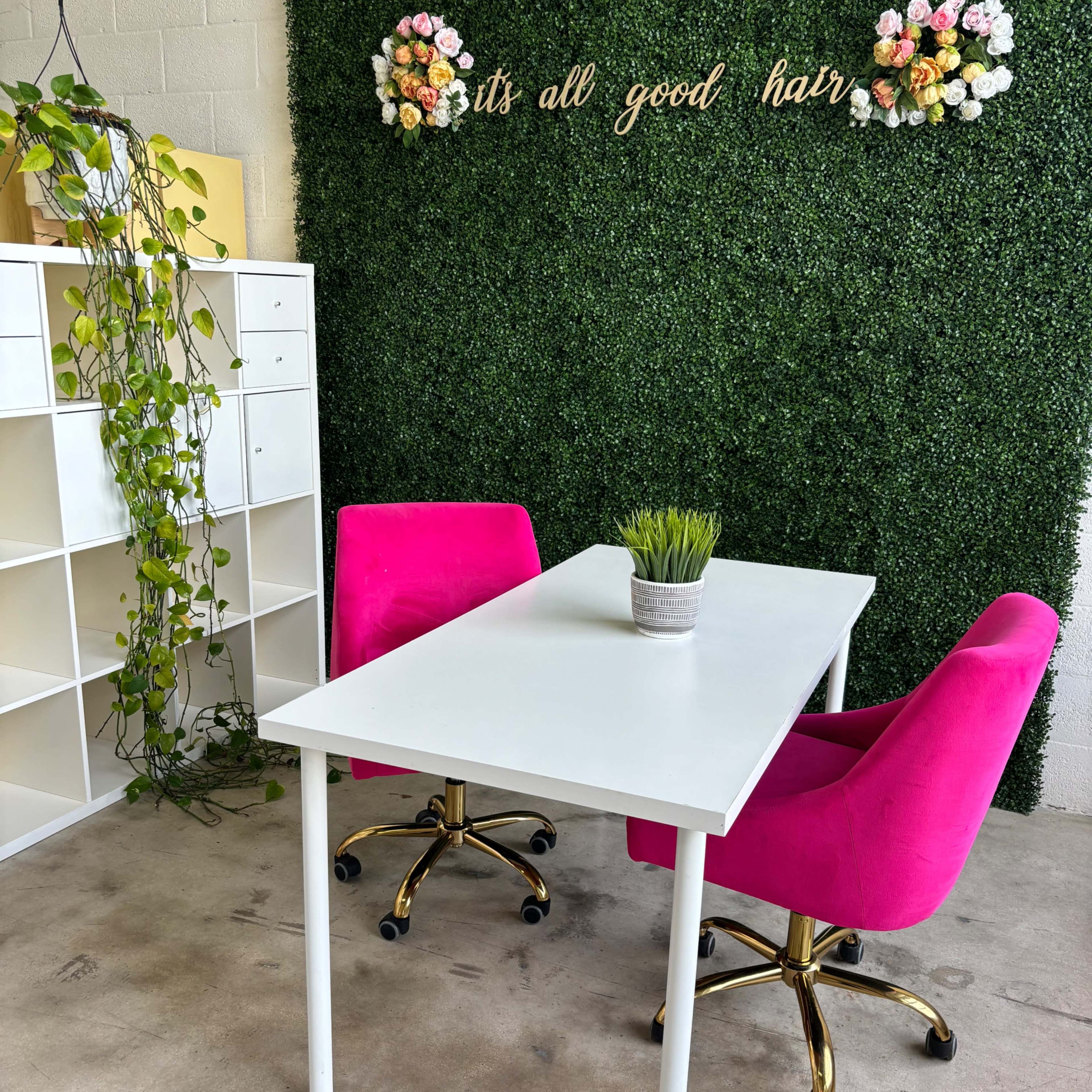 A white table with two pink chairs sits in front of a green wall adorned with flowers and the text "it's all good hair."