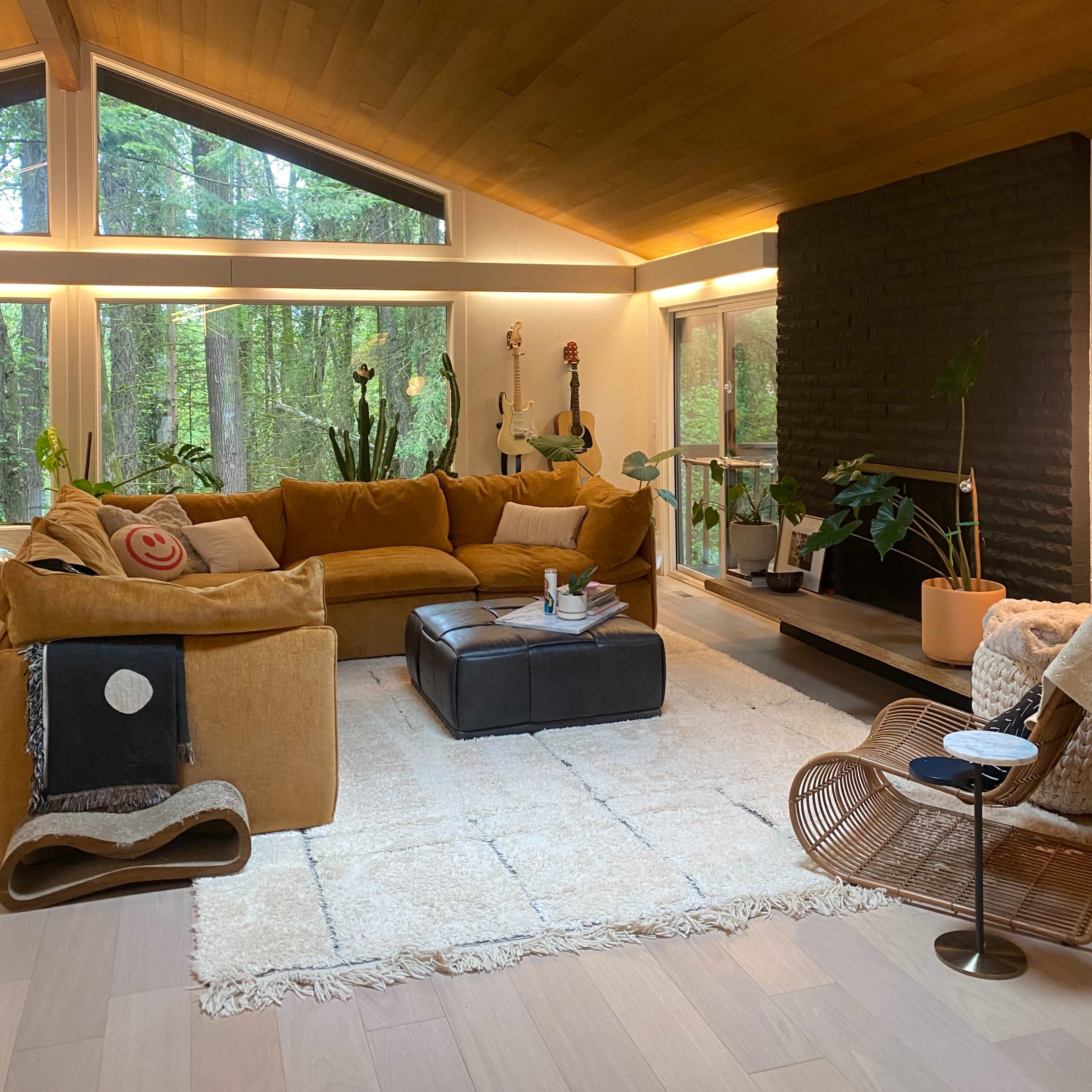 The living room features a large sectional sofa, a central ottoman, and a fireplace, all set against a backdrop of large windows overlooking a forest.