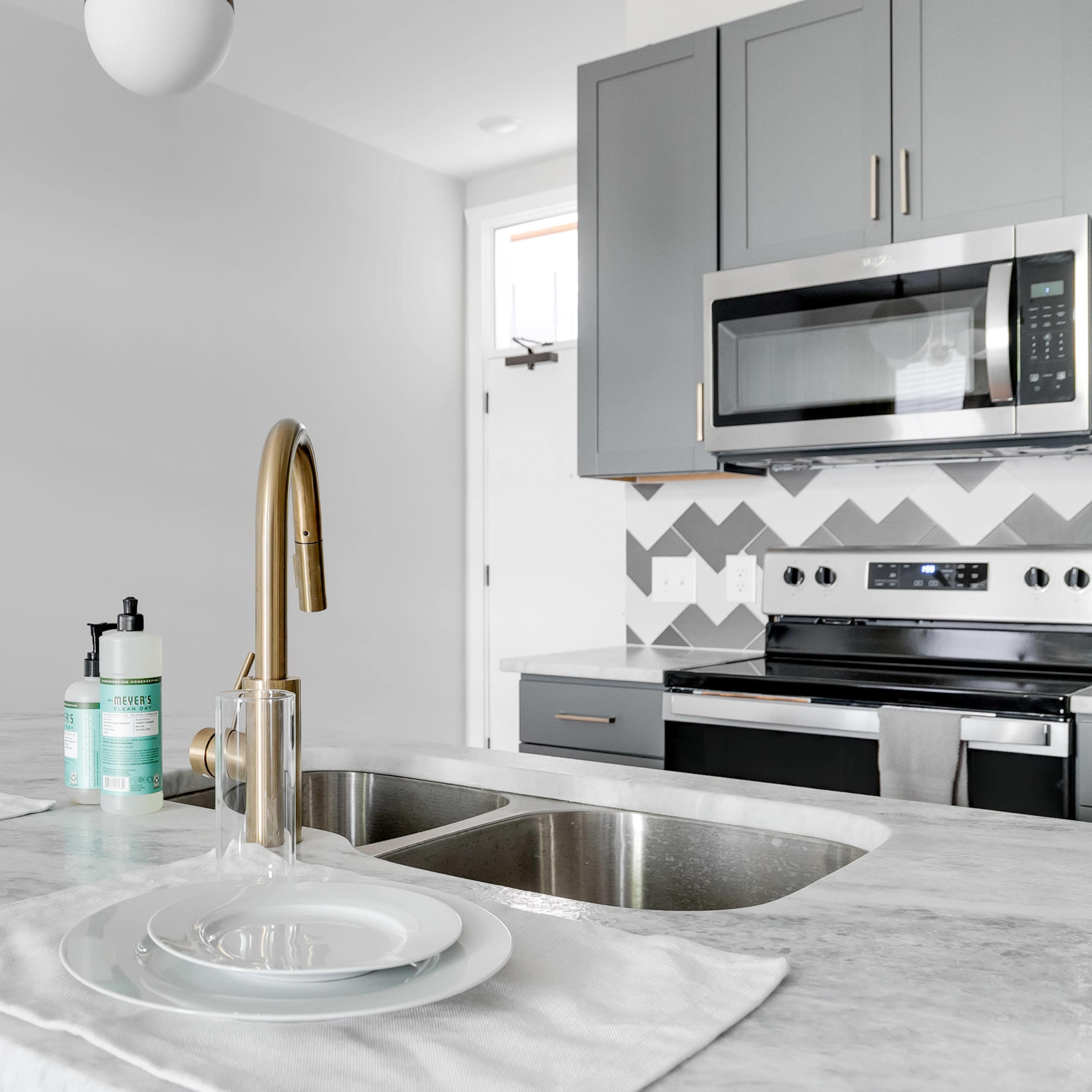 A modern kitchen with a marble countertop, stainless steel appliances, a double sink, and gray cabinets, complemented by chevron patterned tiles on the backsplash.