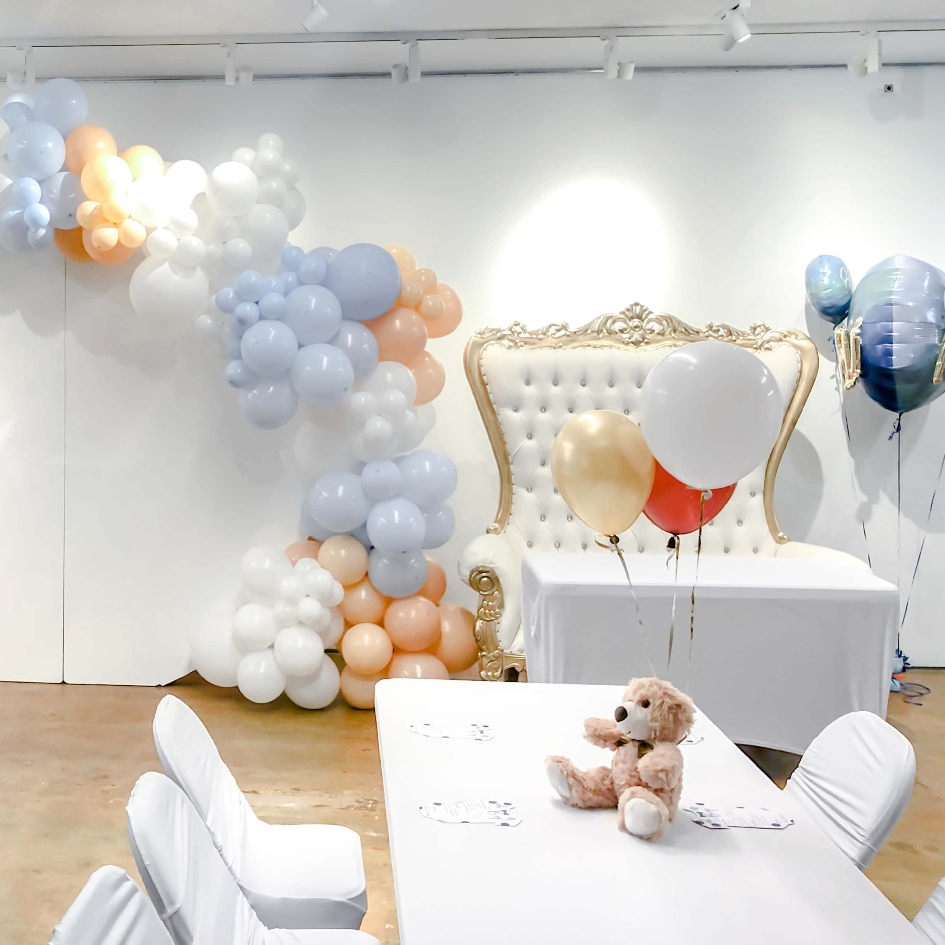 A party venue decorated with a large teddy bear, balloon arrangements, elegant seating, and white tables set for an event.