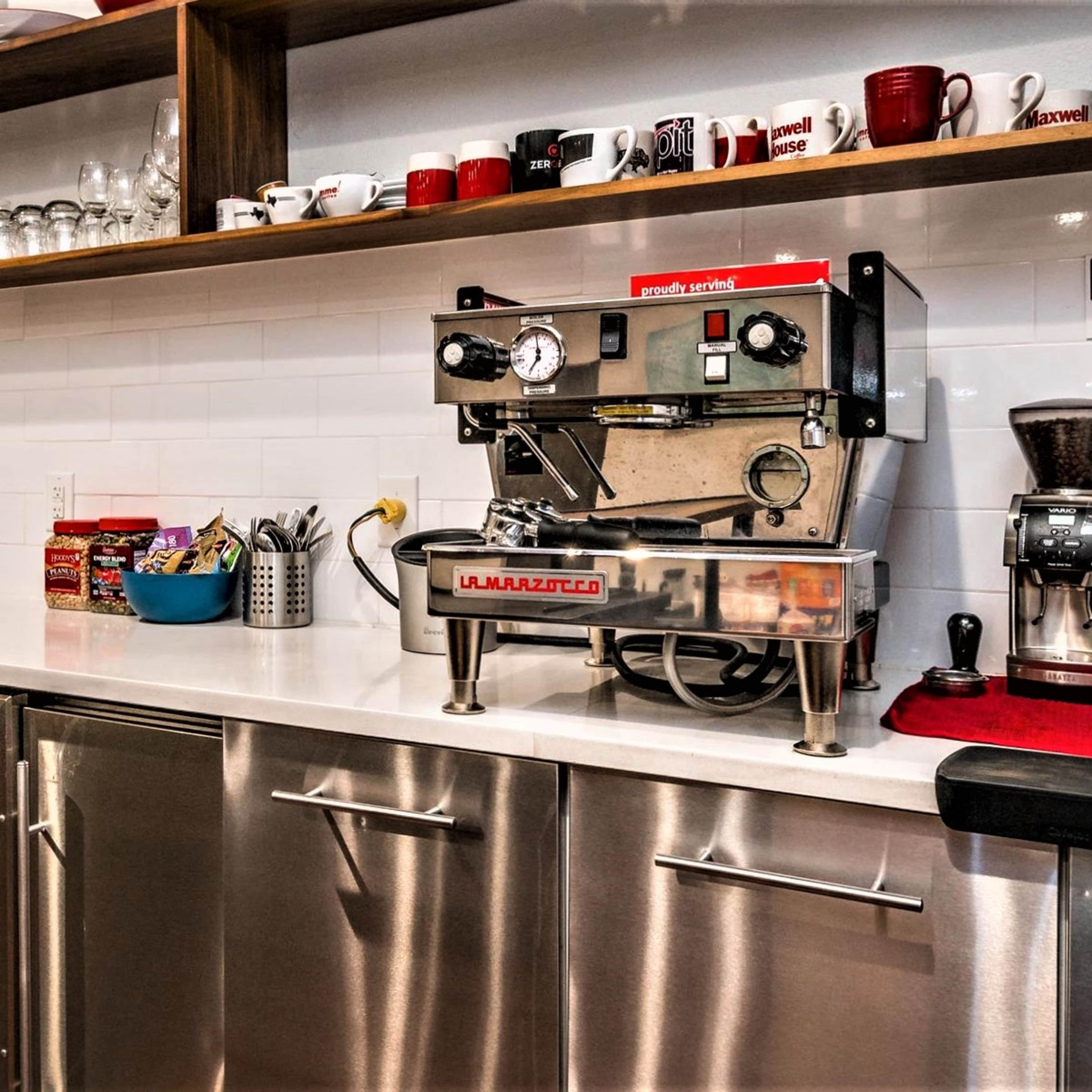 A coffee preparation area featuring an espresso machine, a coffee grinder, glassware, and a wine cooler in a modern kitchen setting.