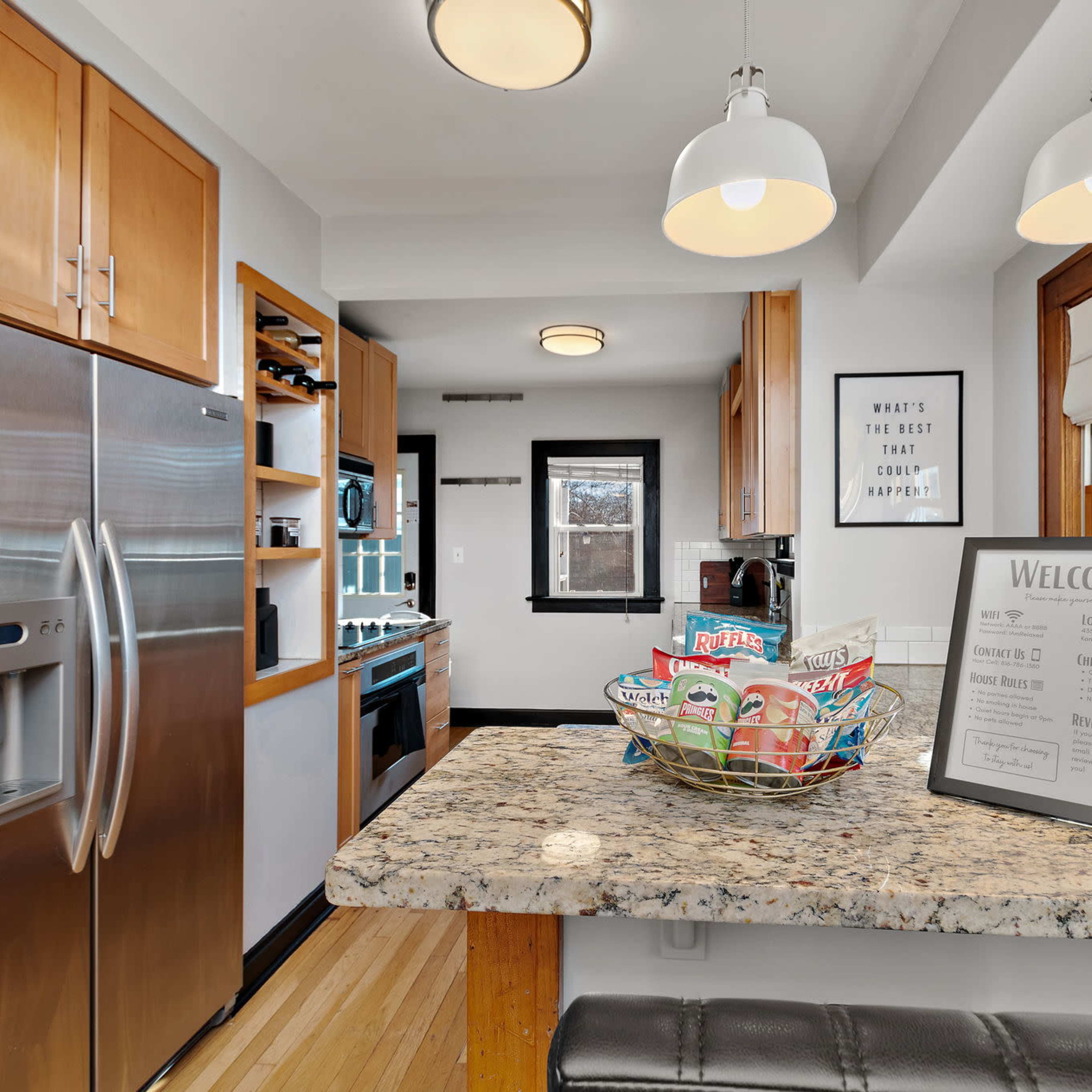 A modern kitchen features stainless steel appliances, wooden cabinets, and a granite countertop with a welcome sign and a colorful basket.