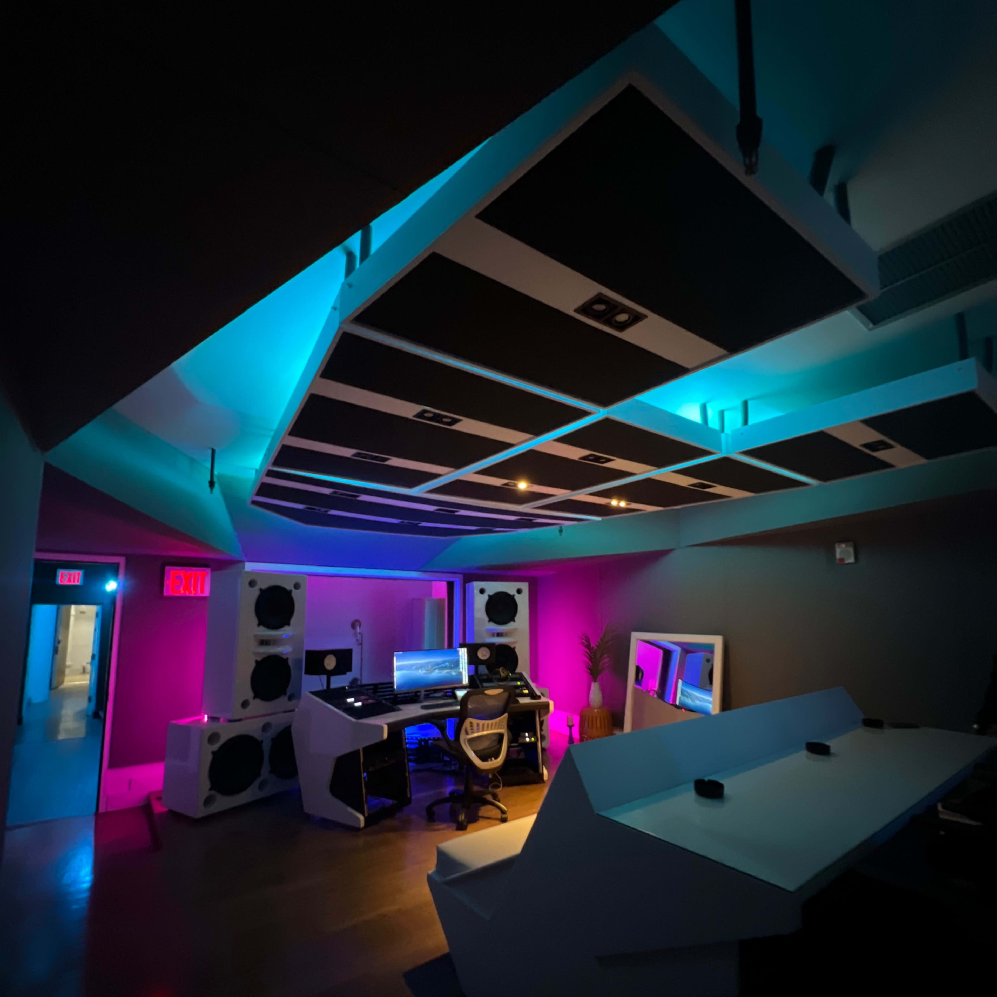 A dimly lit music studio featuring colorful LED lighting, large speakers, and a person working at a computer setup.