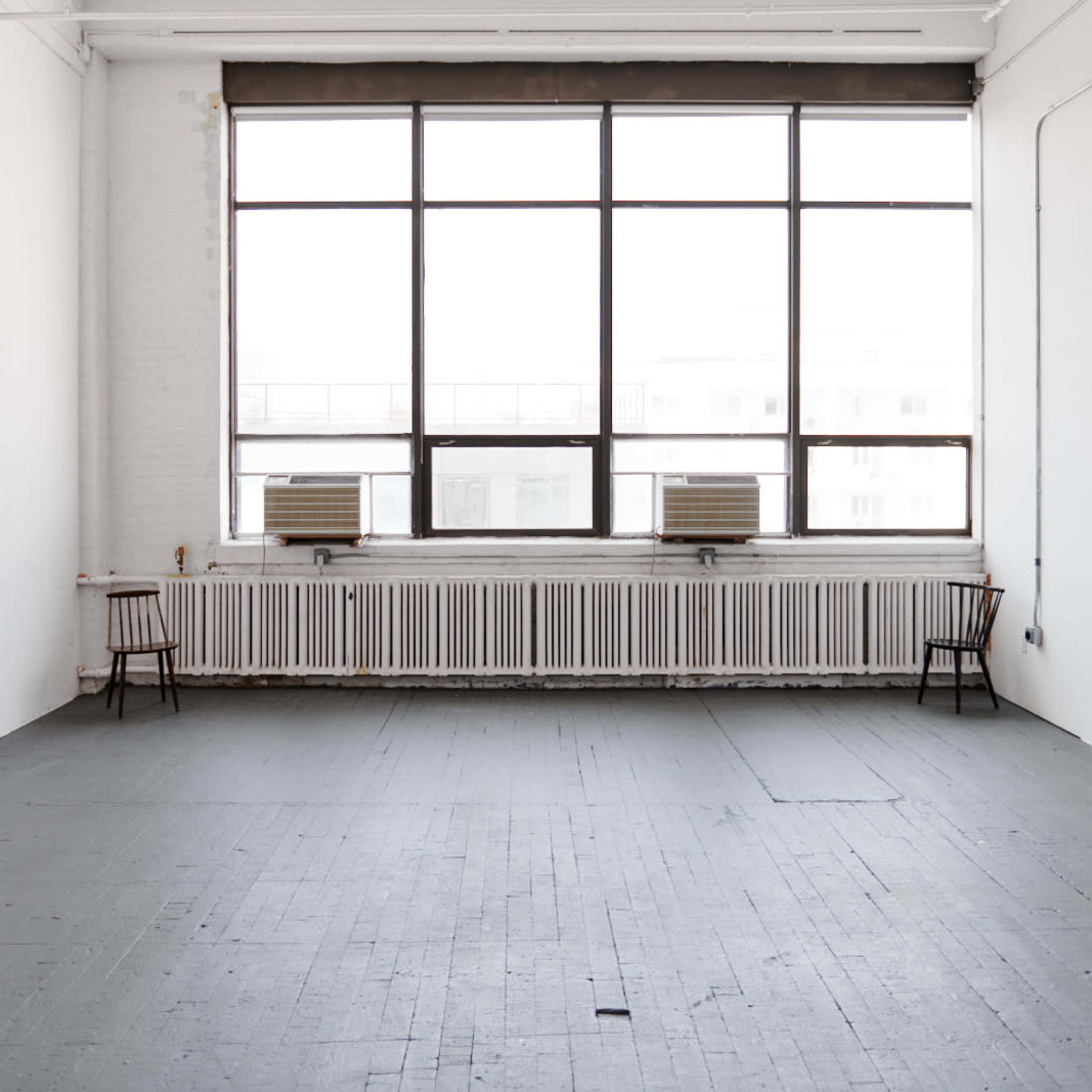 A spacious, empty room with two chairs and a large window, featuring air conditioning units and a radiator along one wall.