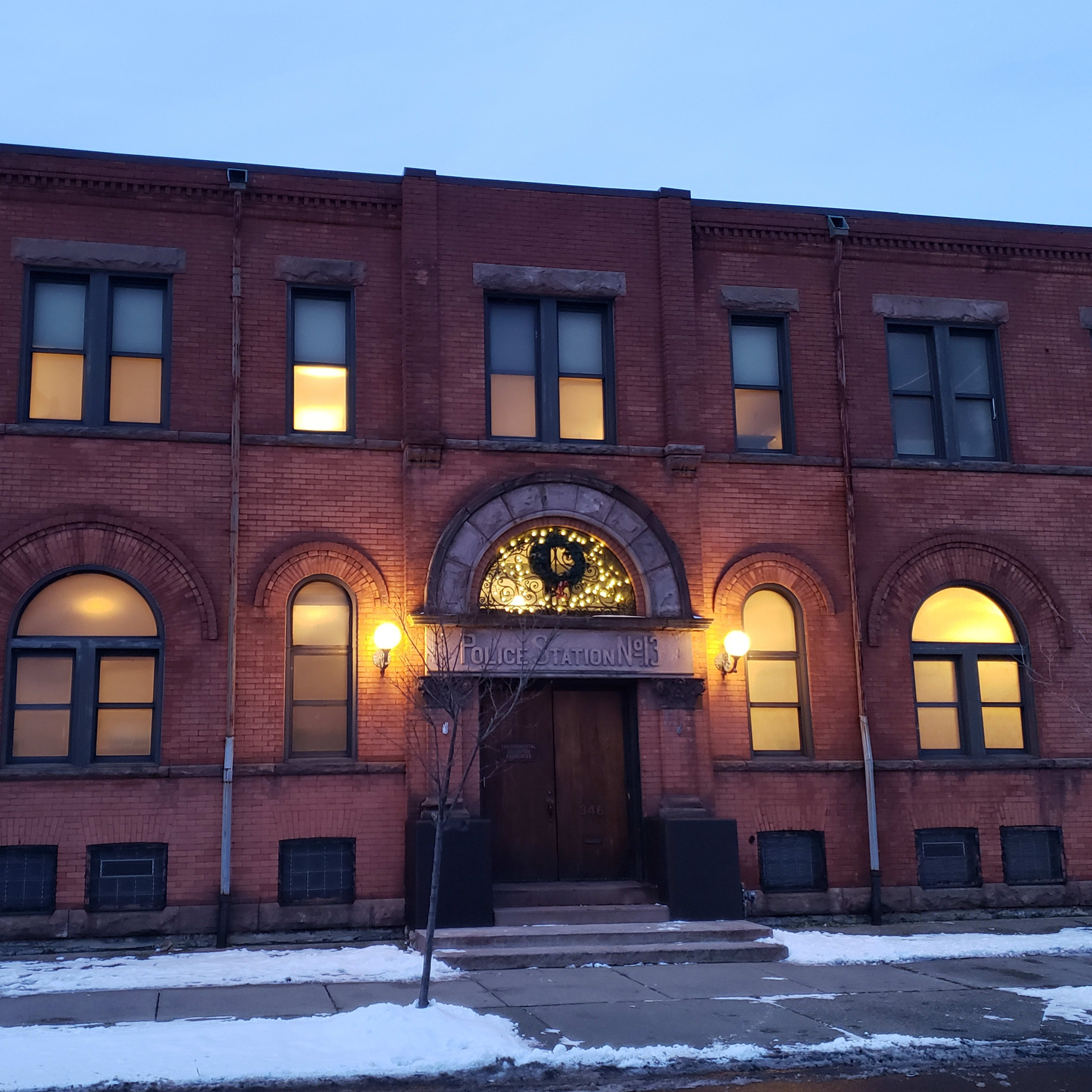 The brick building features arched windows and a decorative clock above the main entrance, illuminated in the evening.