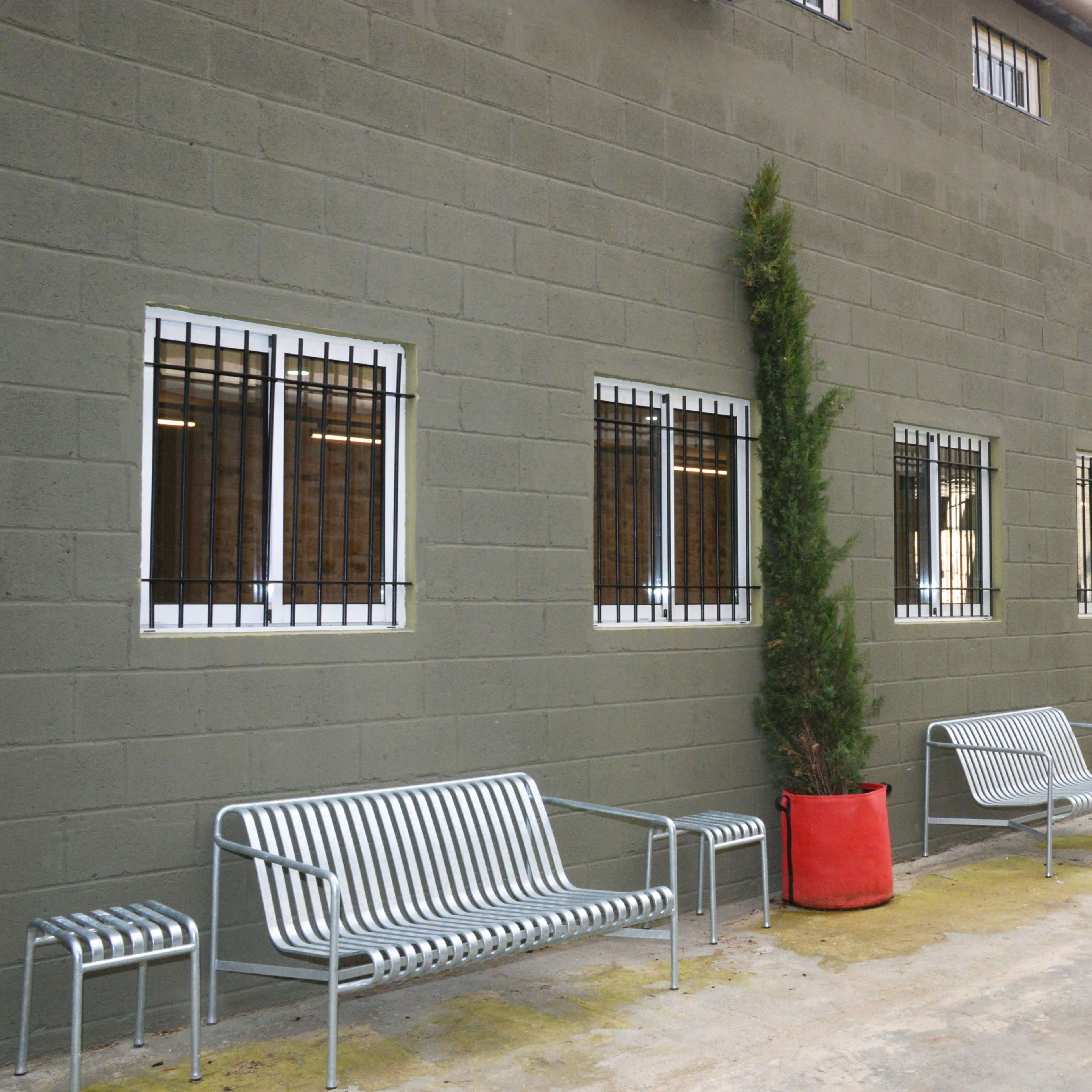 A row of silver benches and potted conifer trees lines the side of a gray building with barred windows.