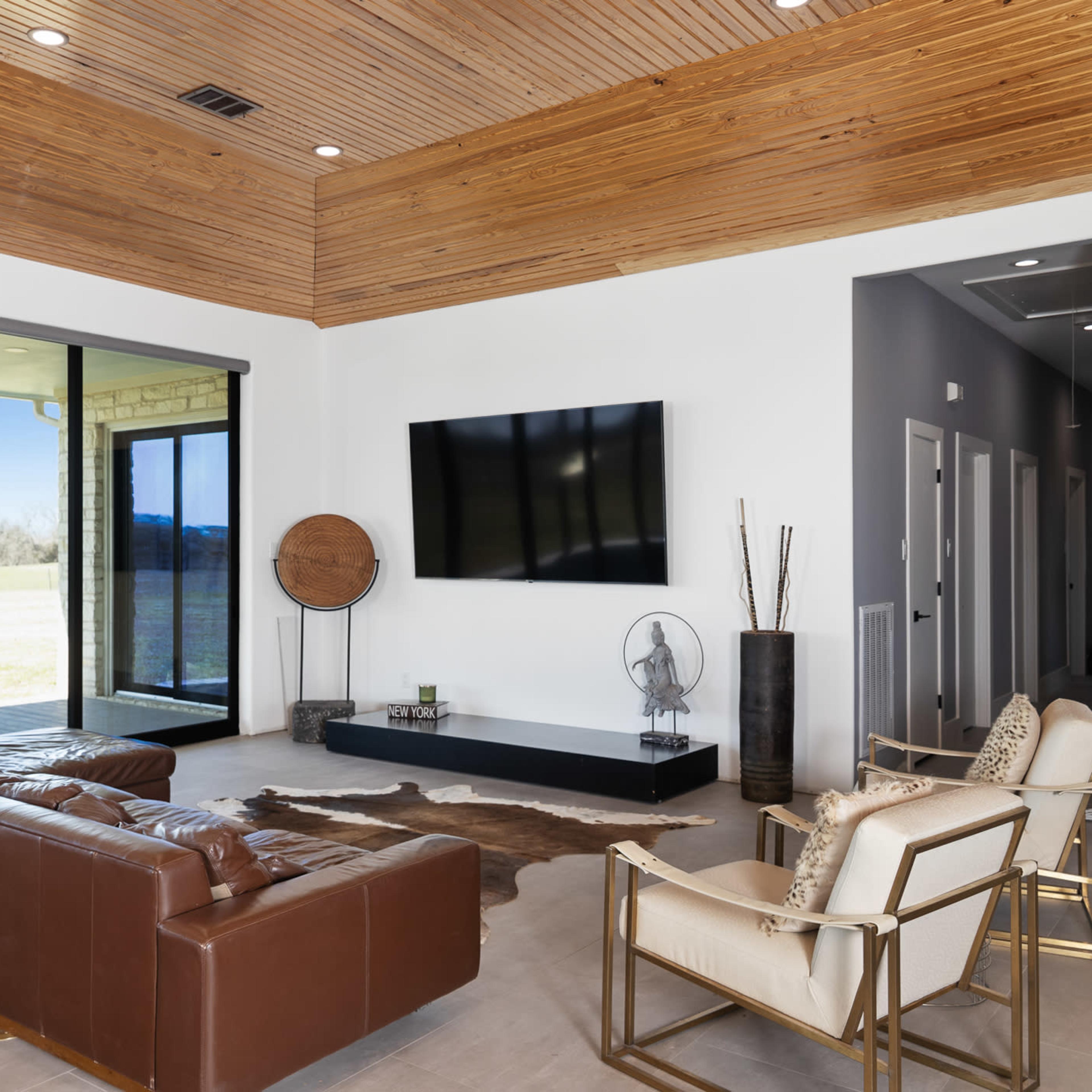 A modern living room features a leather sofa, an accent chair, a wall-mounted television, and large sliding glass doors leading to an outdoor view.