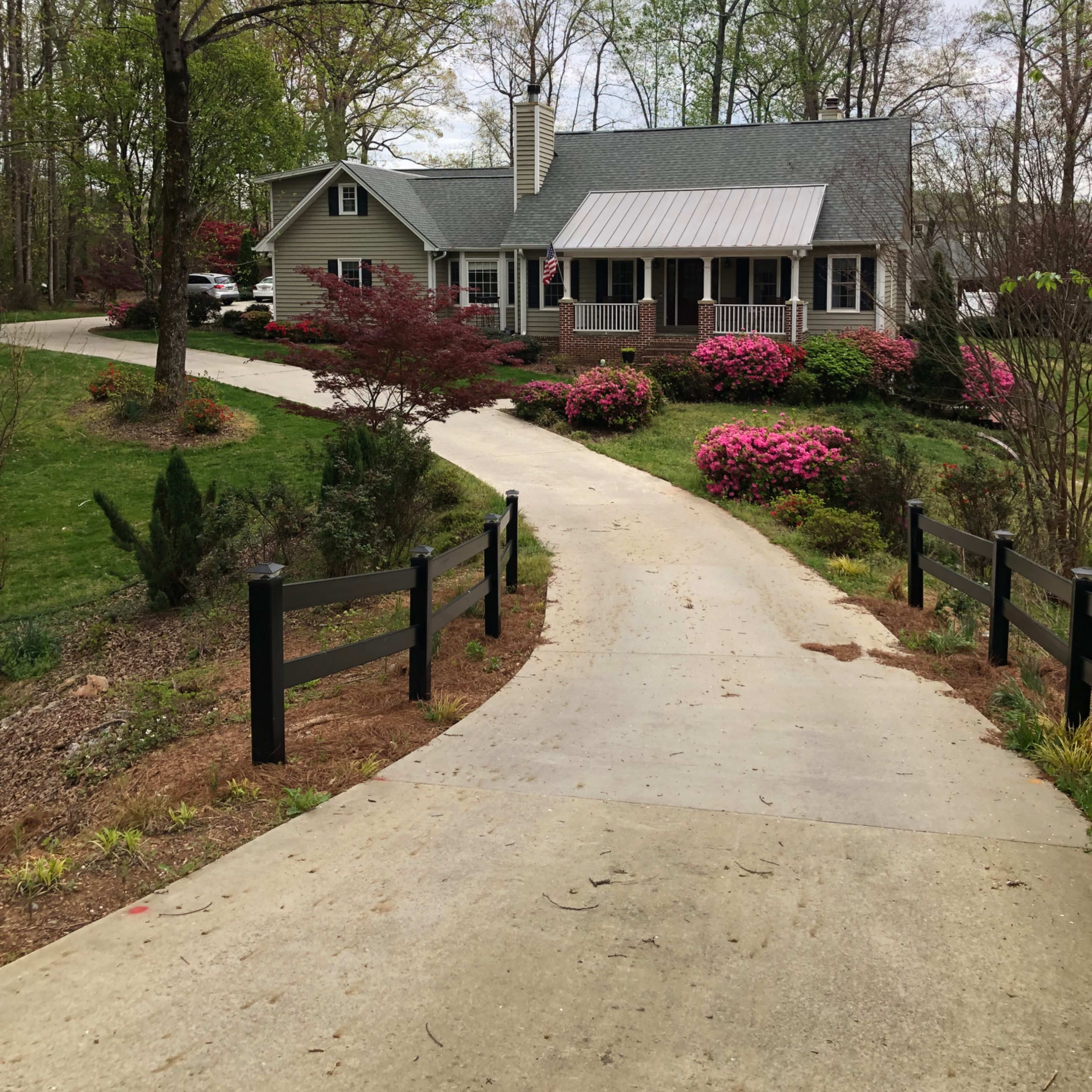 A gravel driveway leads to a single-story house surrounded by colorful flower beds and greenery.