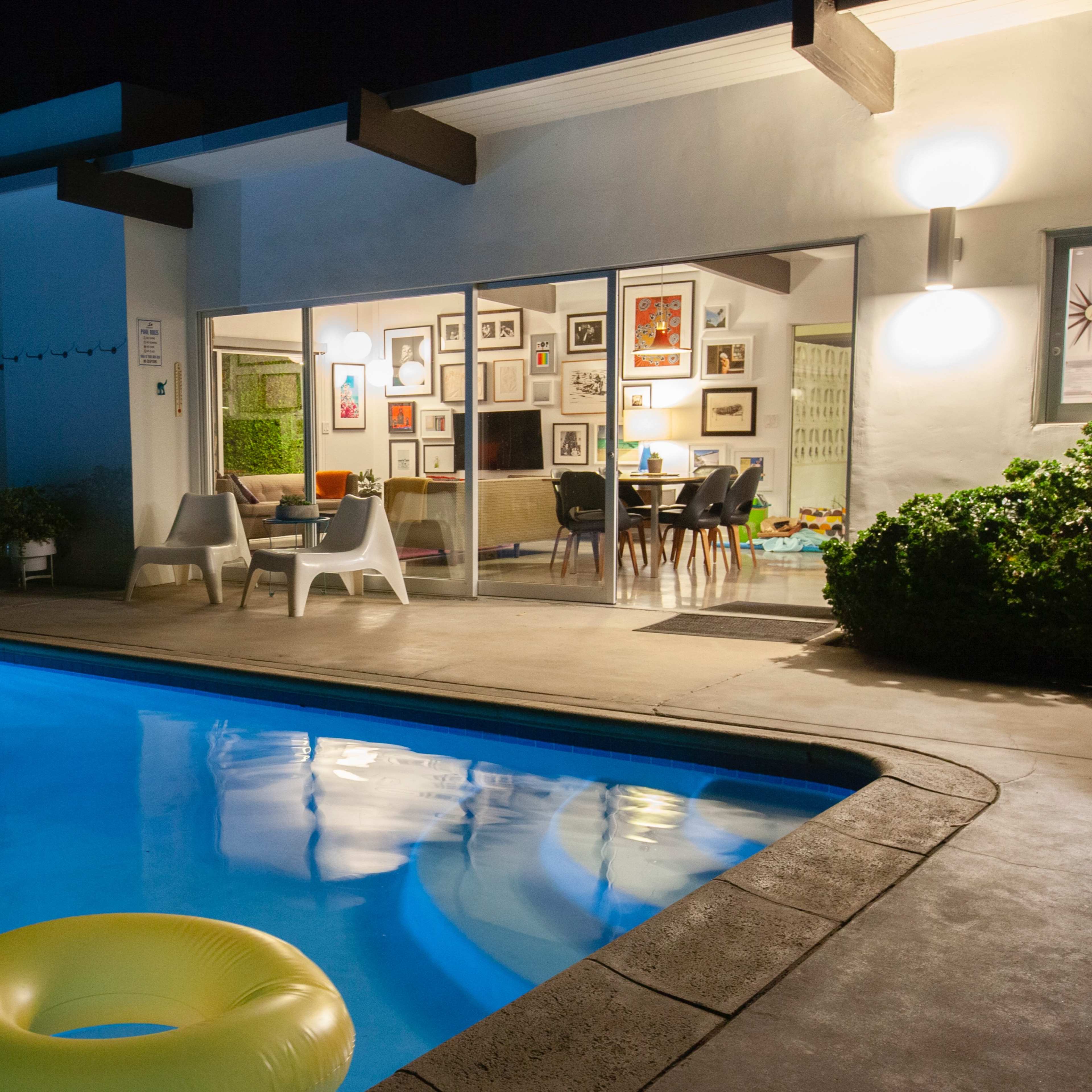 A brightly lit residential backyard at night features a swimming pool with a yellow float, surrounded by greenery and a house with framed pictures on the walls.