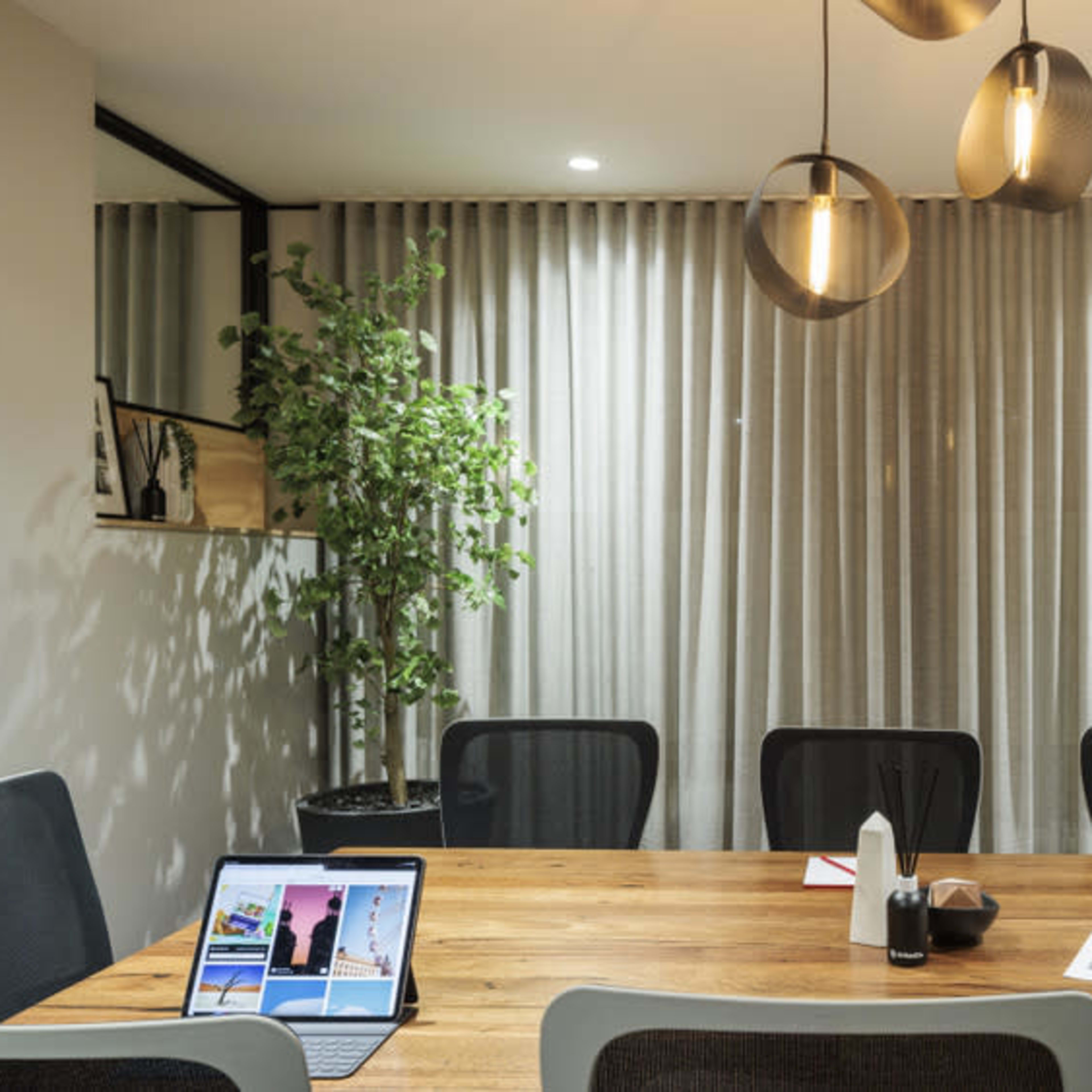 A modern conference room features a large wooden table surrounded by eight chairs, with a television on the wall and a plant in the corner.