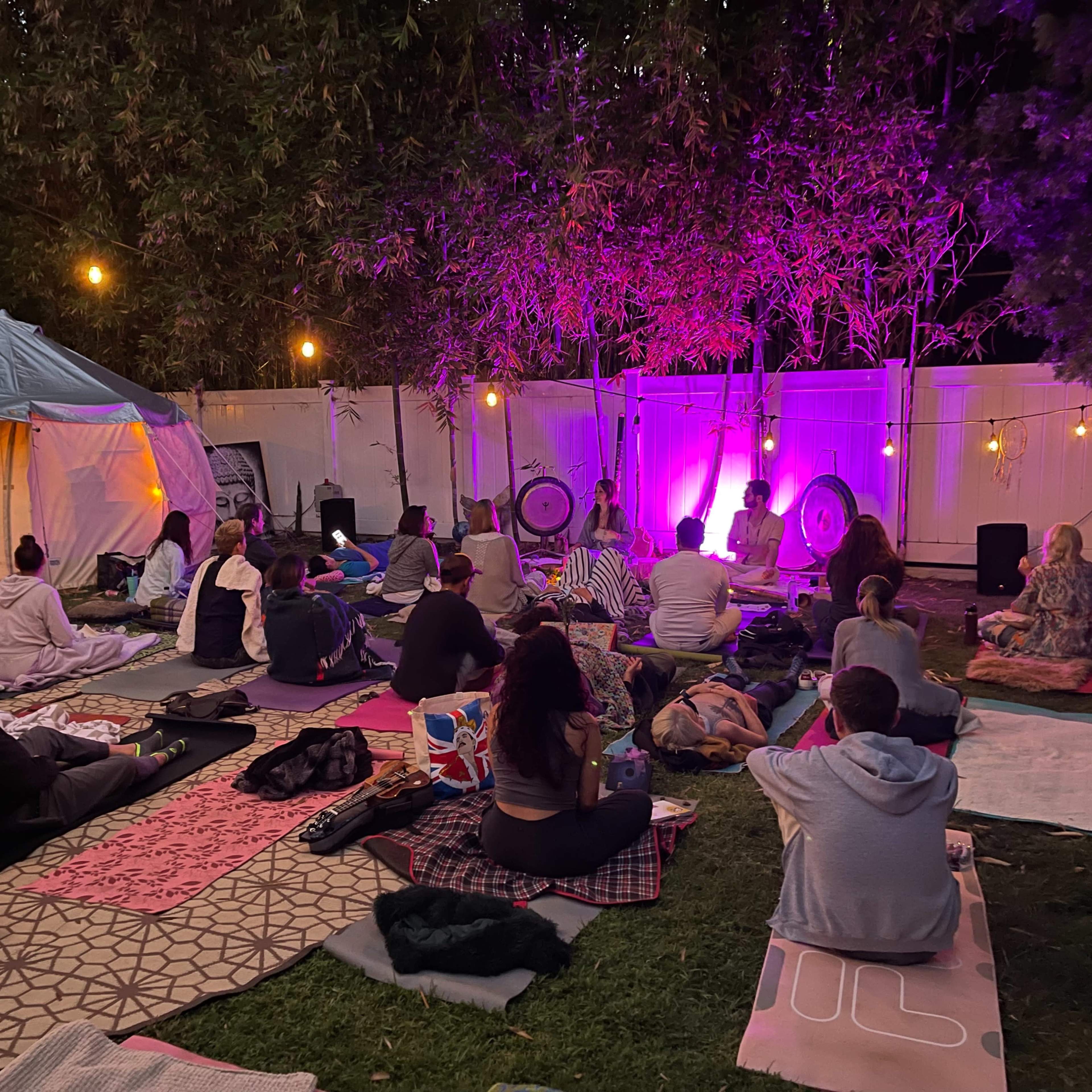 A group of people sits on blankets in a garden setting, facing a stage with purple lighting and musical instruments.