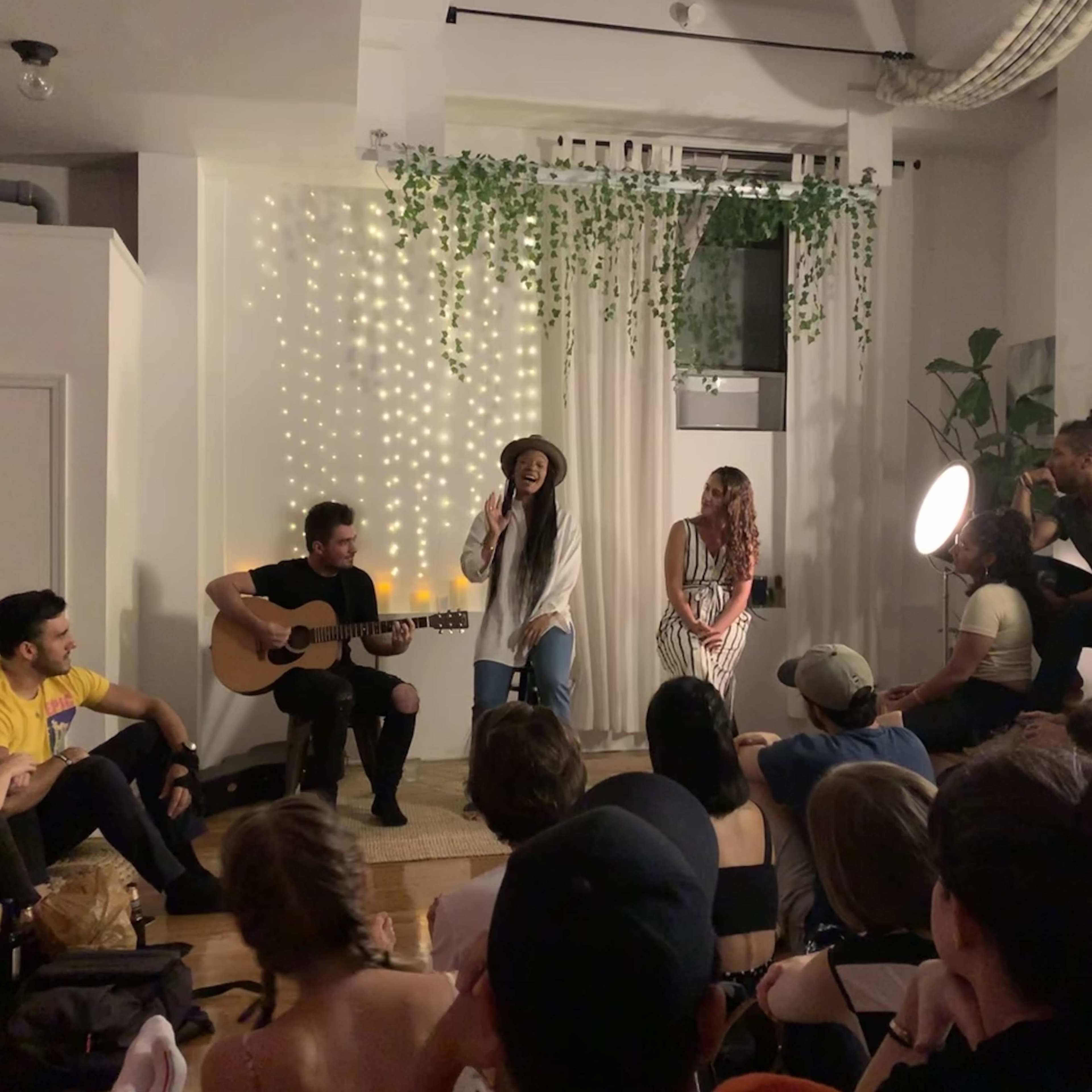 A singer performs on stage with an acoustic guitarist in front of an attentive audience in a stylized indoor setting.