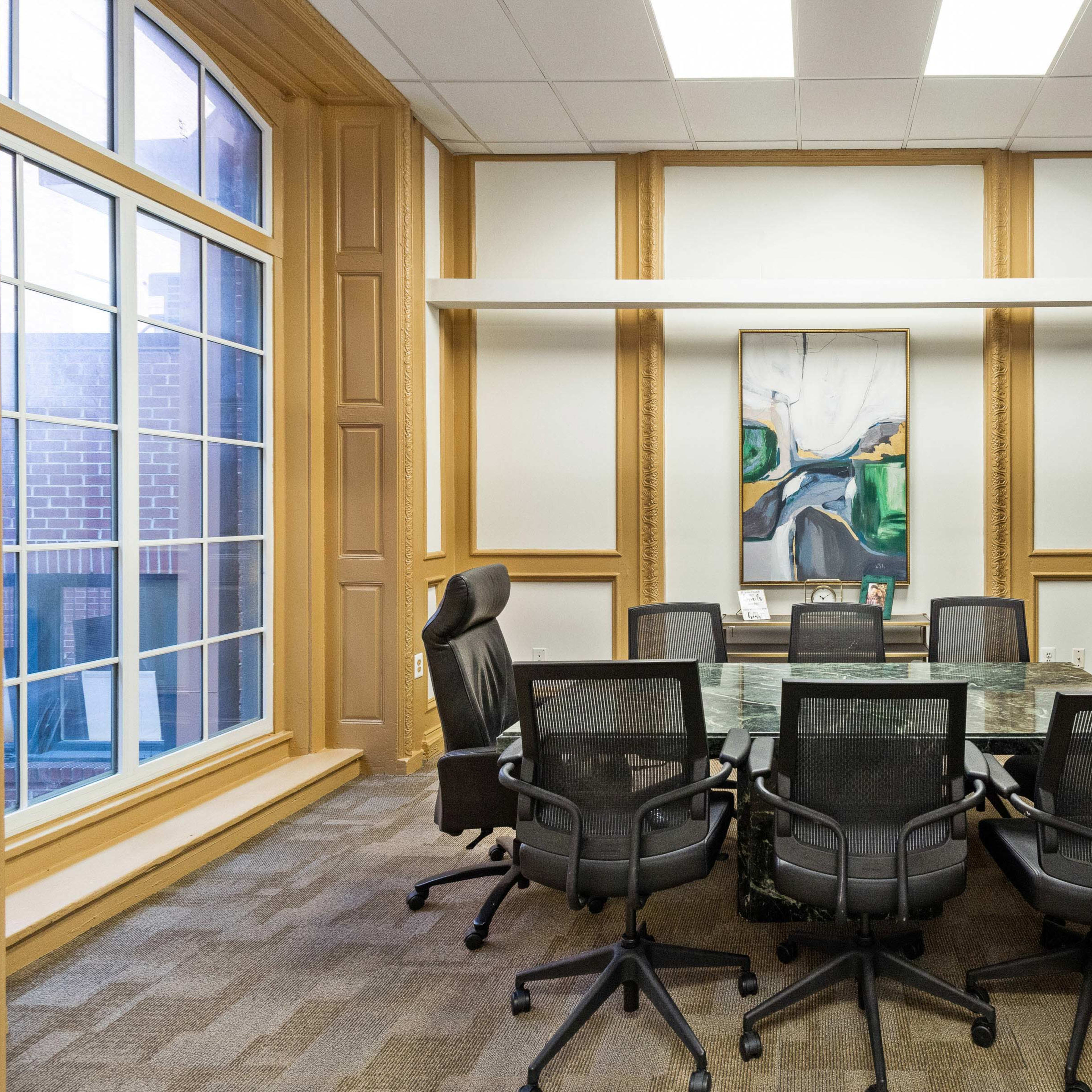 A large conference room features a glass table surrounded by black mesh chairs, with windows on one wall and a large artwork hanging on another.