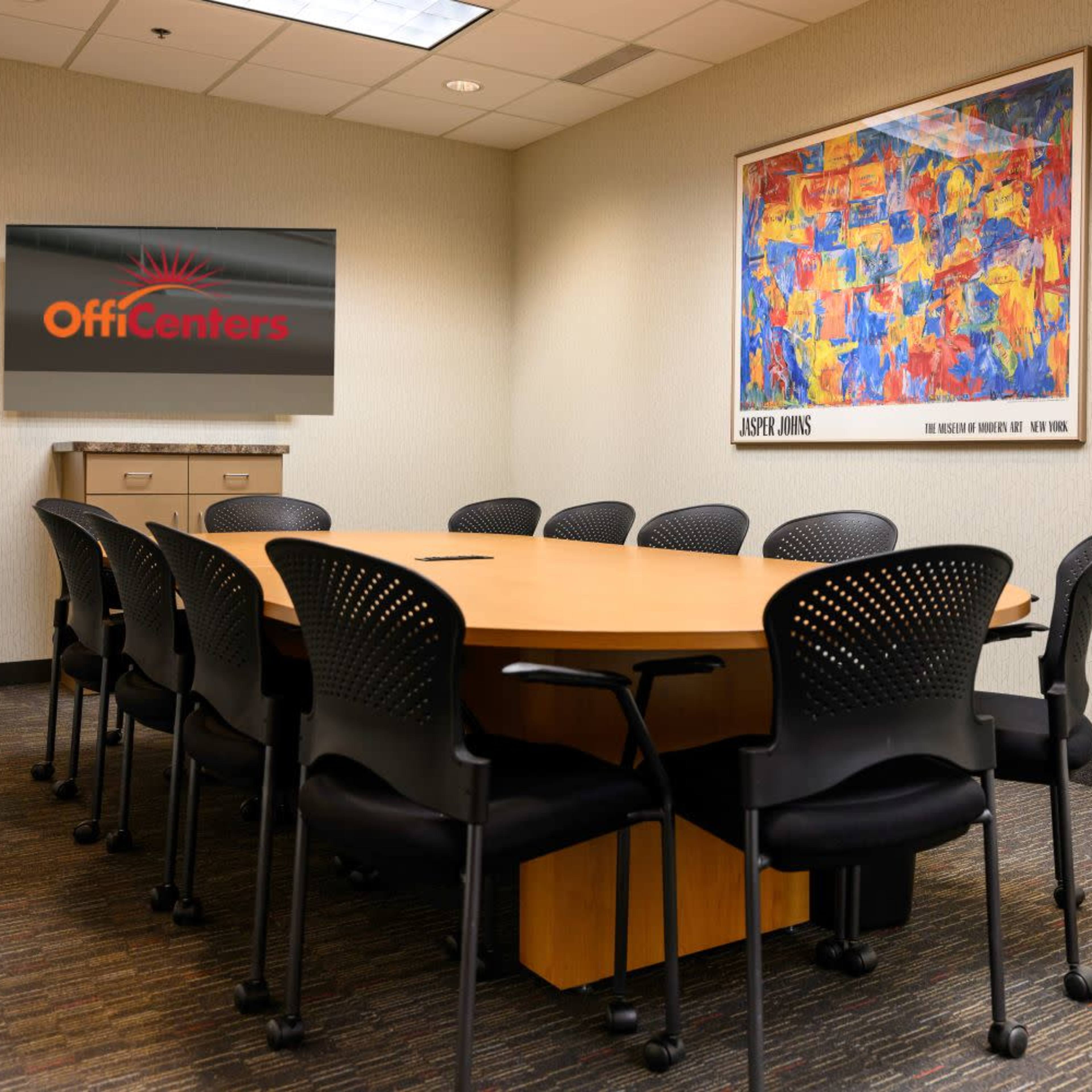 A conference room features a large oval table surrounded by black chairs and a wall-mounted screen displaying the logo "OffiCenters" alongside an abstract artwork.