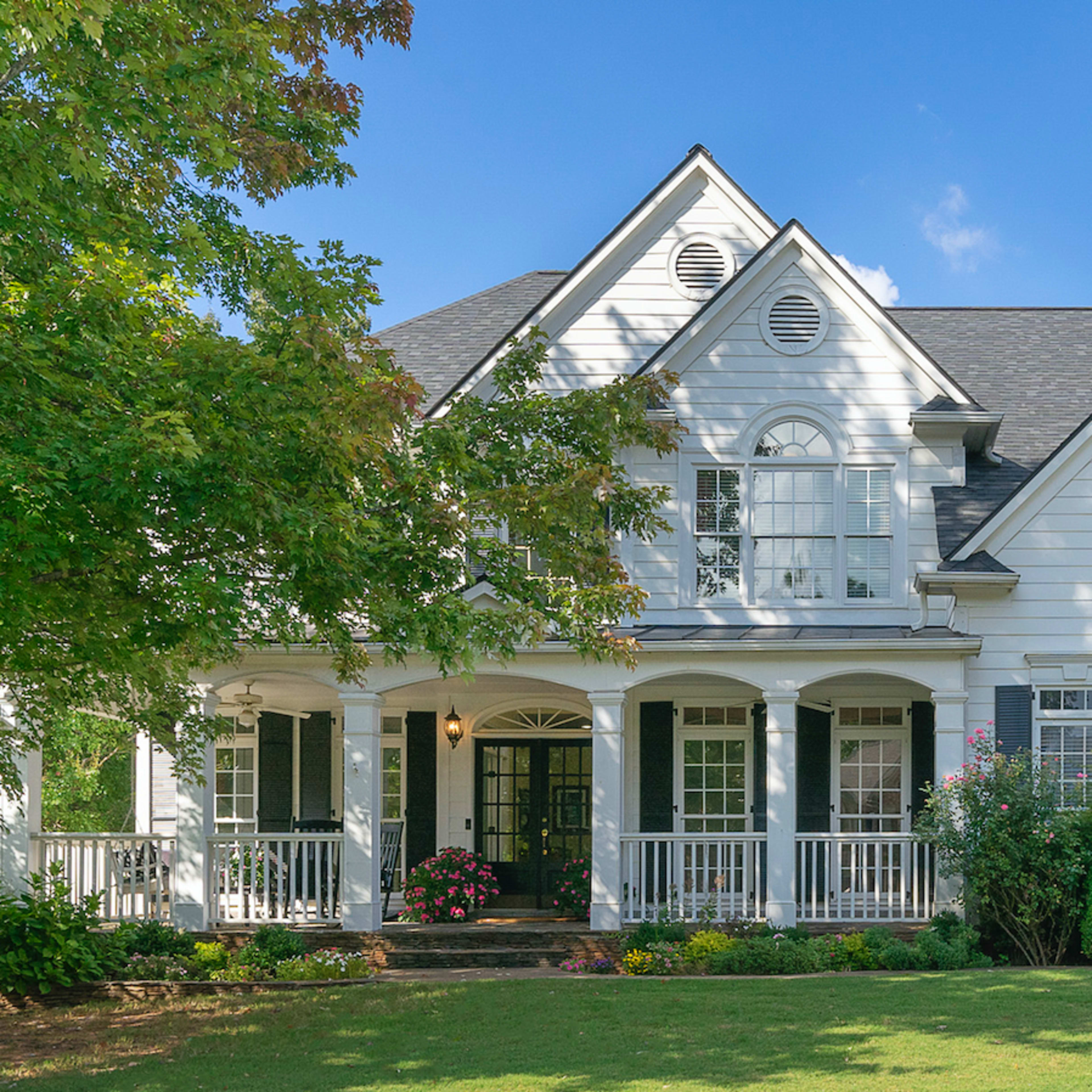A large, two-story white house with a front porch is surrounded by greenery and has multiple windows.