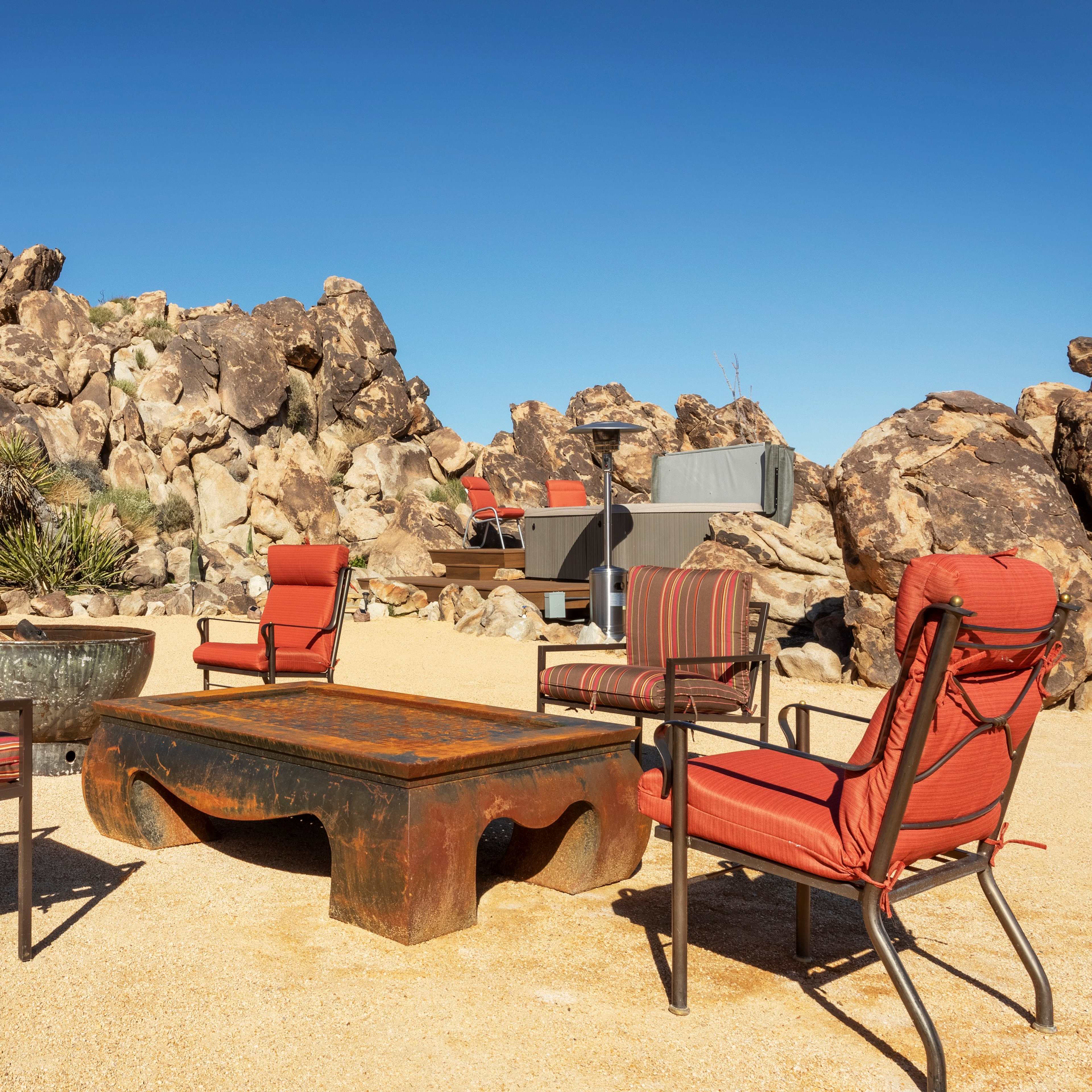 A seating area with metal chairs and a table is set up on a sandy surface near large rocky formations under a clear blue sky.