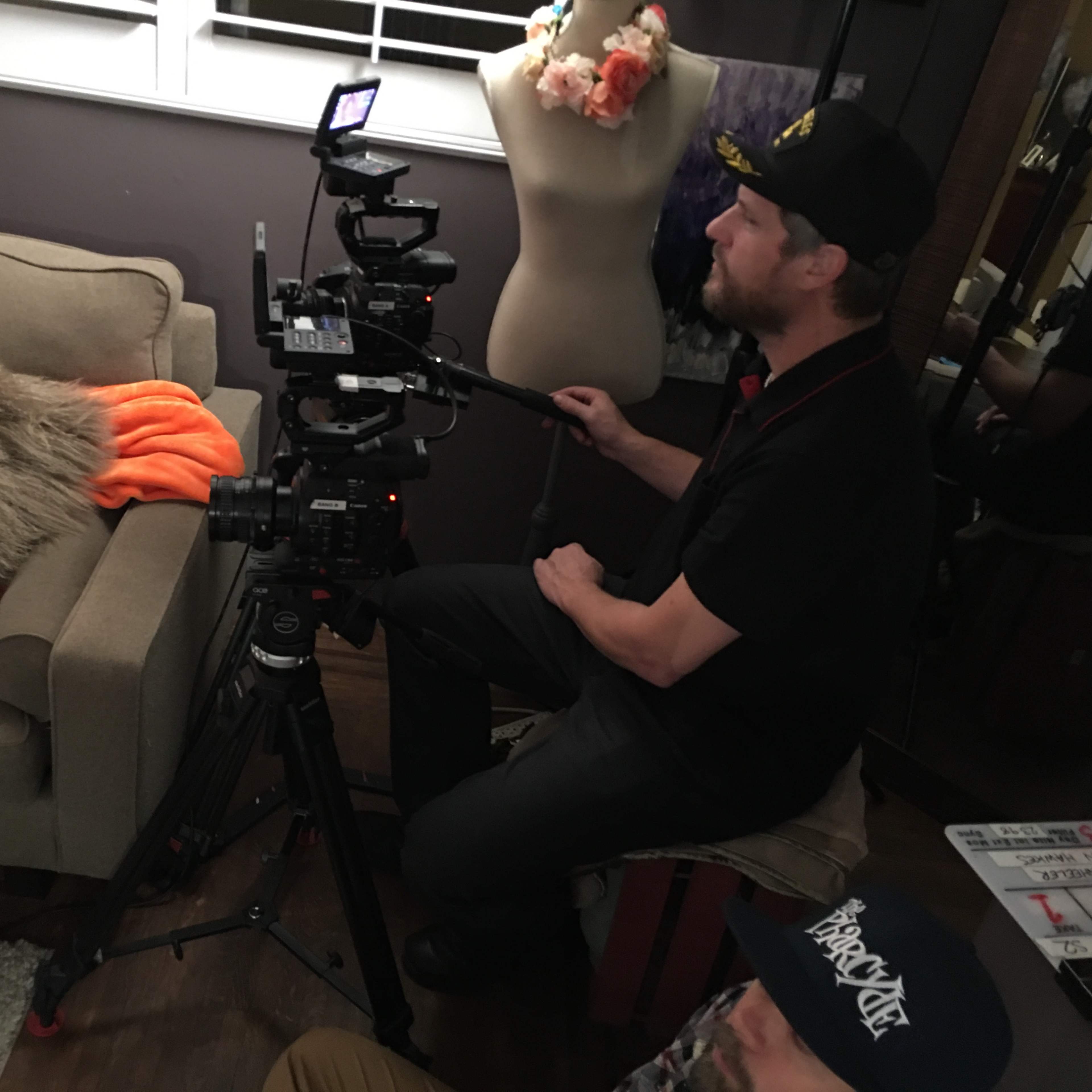 A cameraman operates a professional camera on a tripod while sitting on a stool in a room with a couch and a dress form.