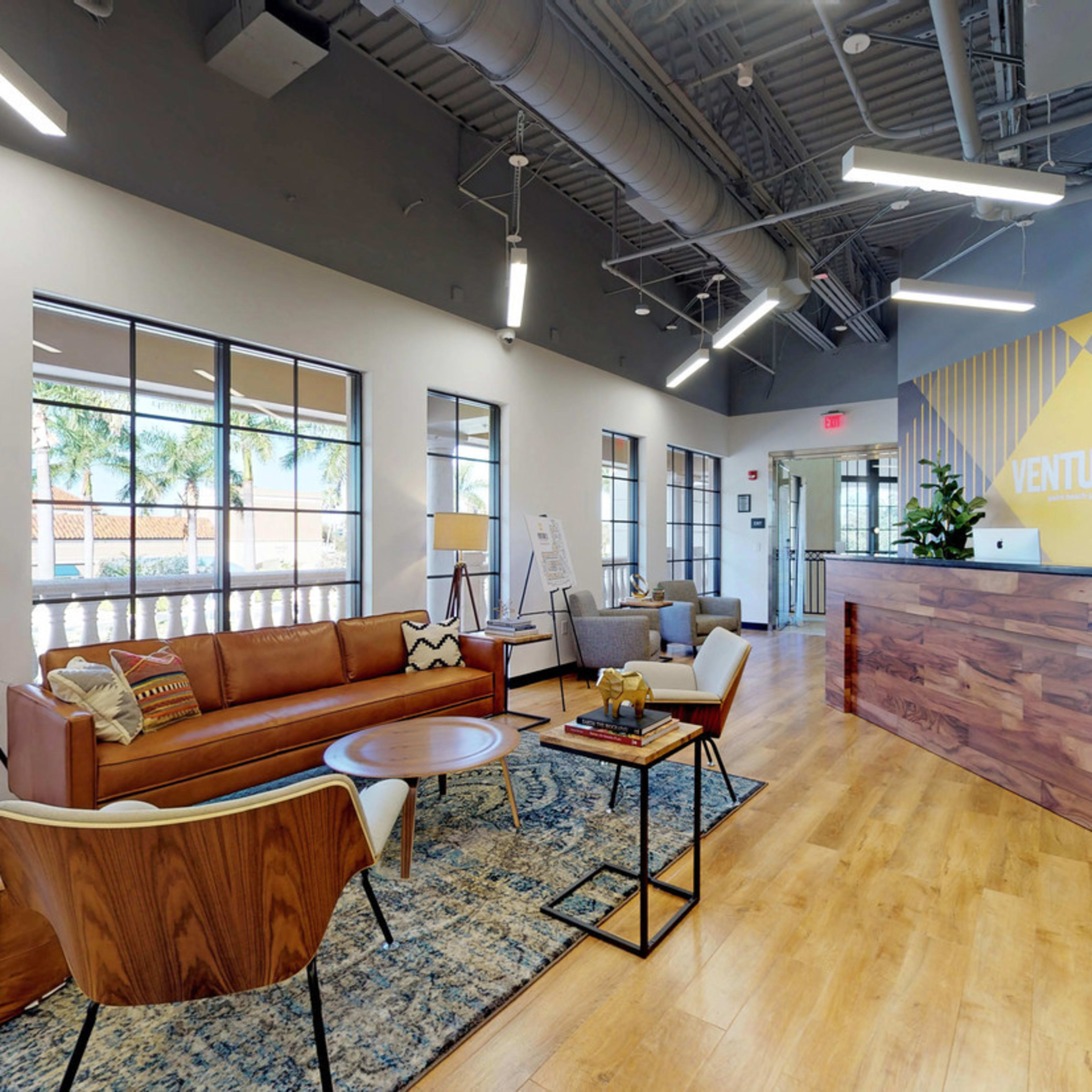 A modern reception area featuring wooden furniture, a large desk with the text "VENTURE X," and a seating arrangement with various chairs and a coffee table.