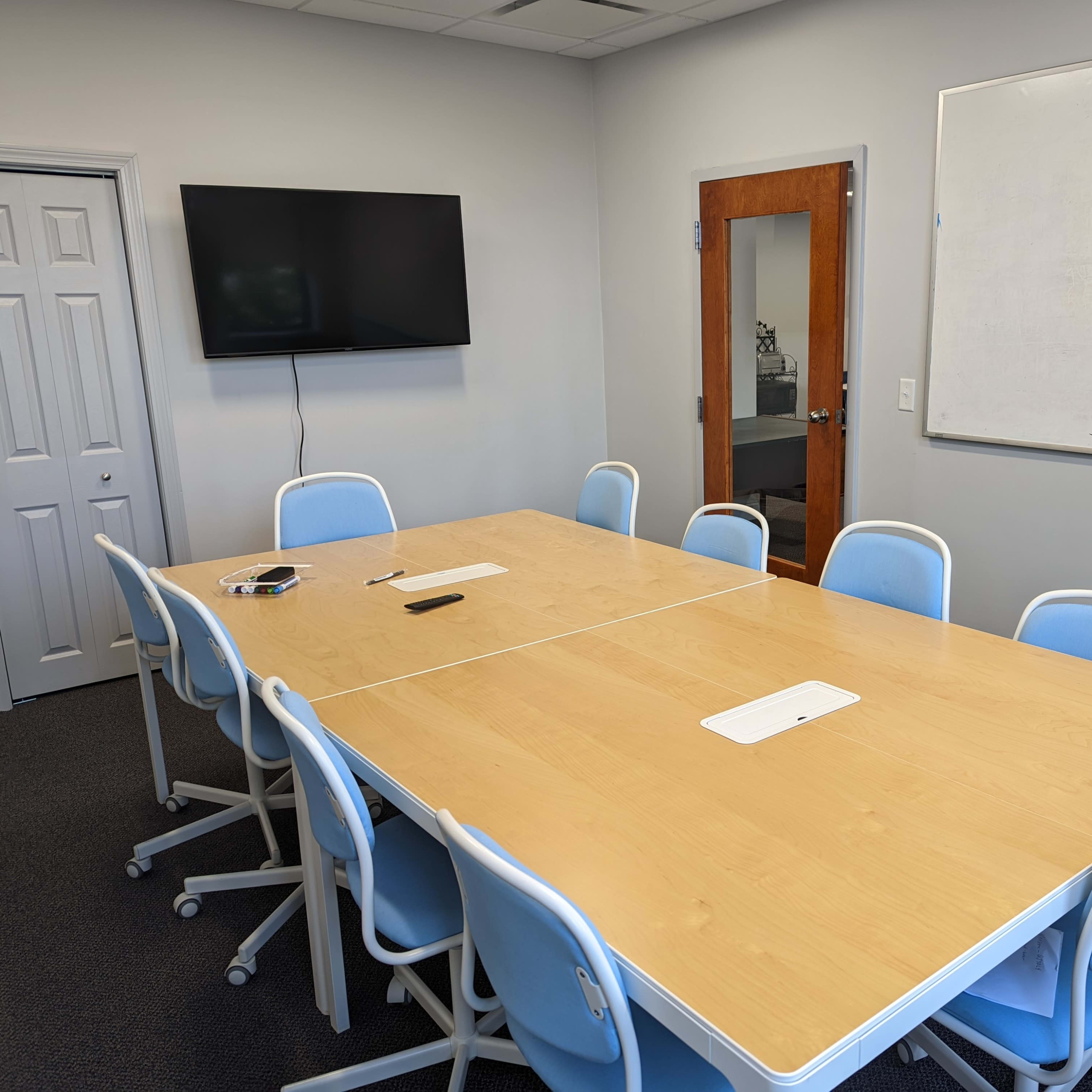 A long wooden conference table with ten blue chairs is positioned in a meeting room, featuring a television on the wall and a whiteboard nearby.
